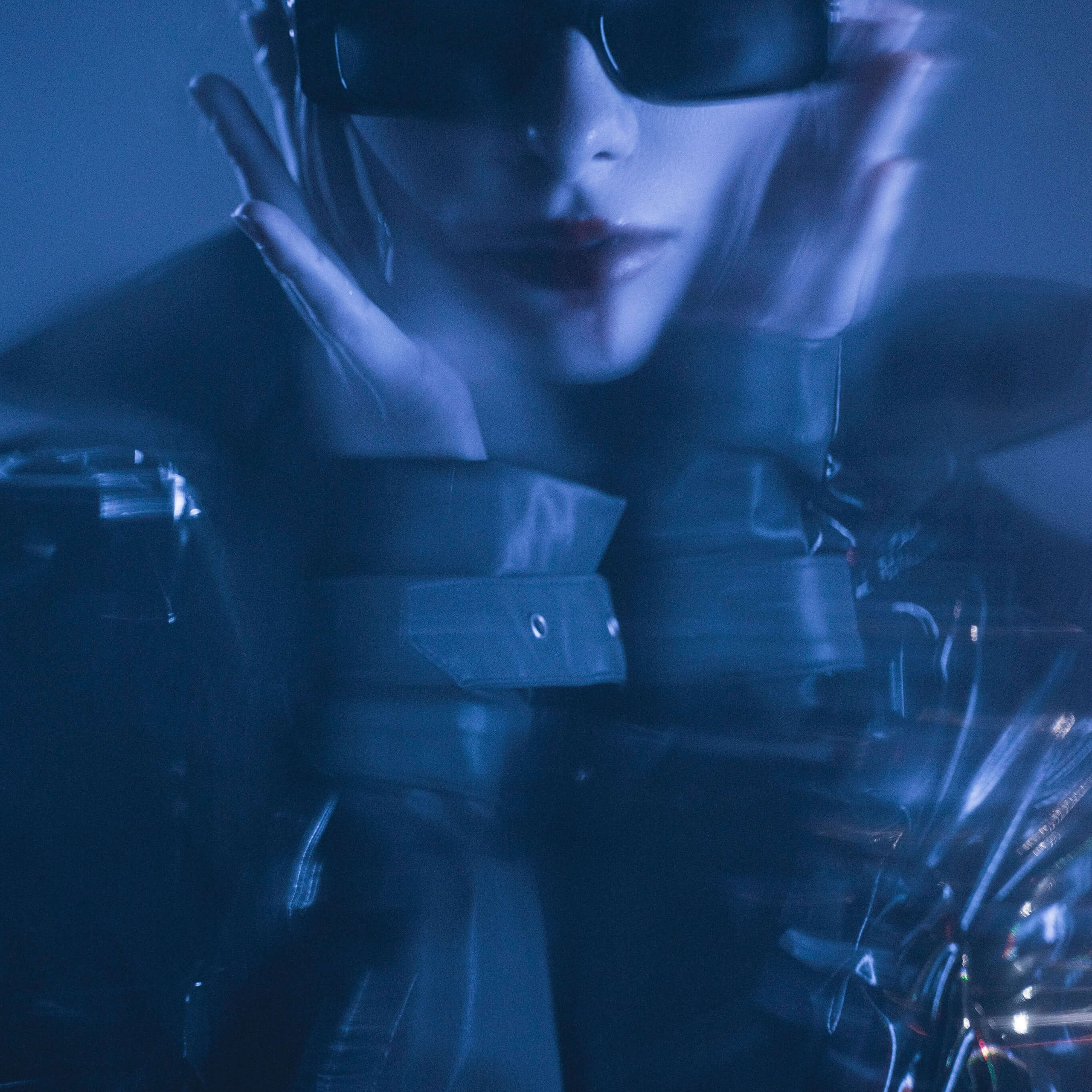 A moody, ethereal portrait of a person in sleek sunglasses and glossy attire, shrouded in a cool blue haze that creates a mysterious vibe