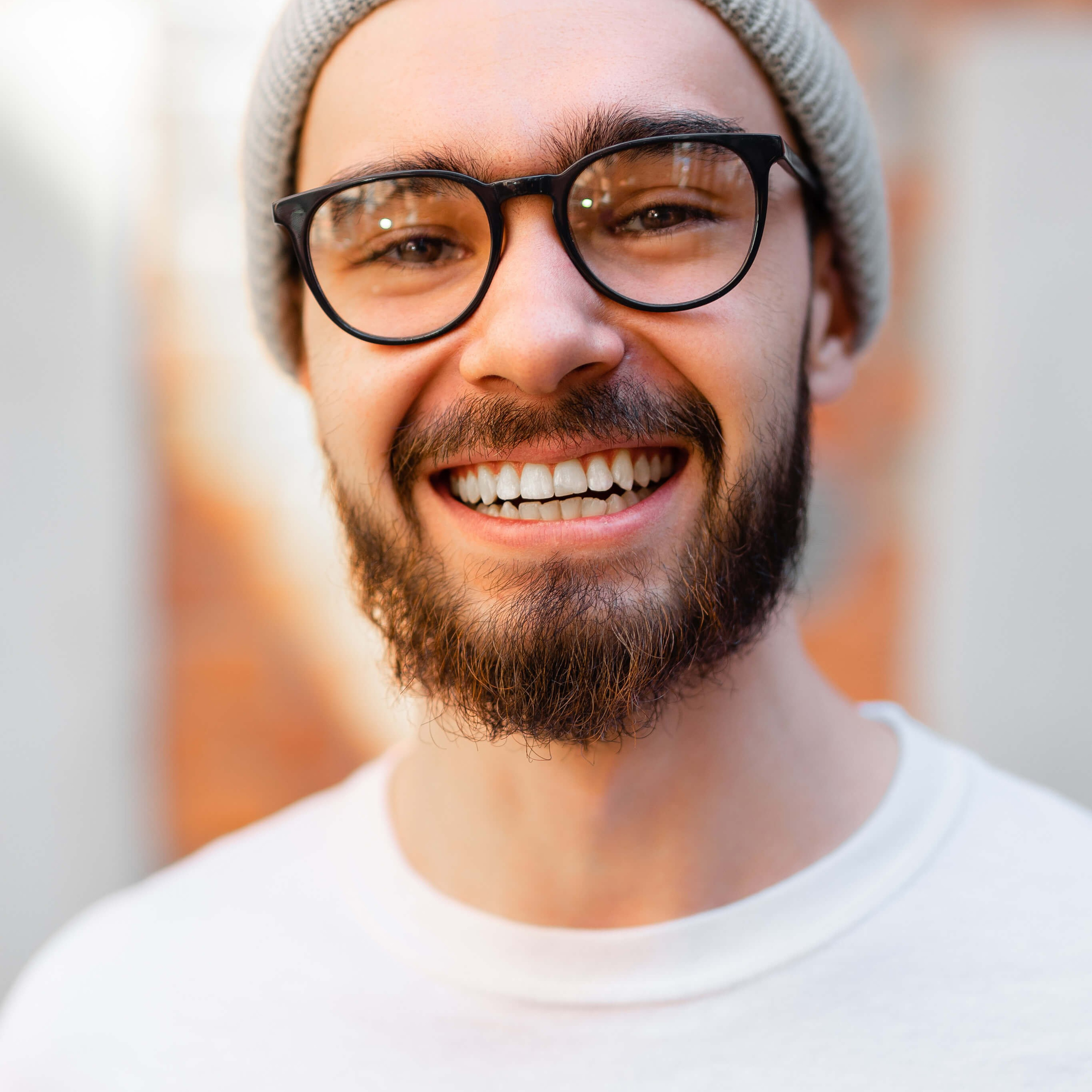 A warm and friendly portrait of a man with a beanie and glasses, smiling broadly, set against a blurred background with soft lighting that highlights his cheerful expression