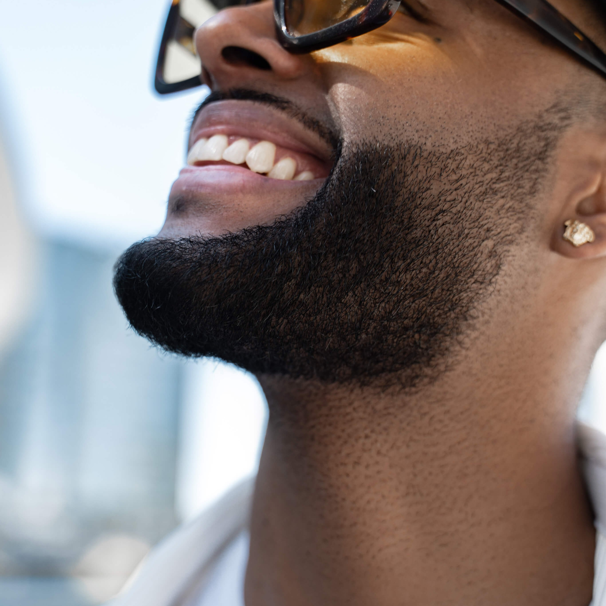 Close-up of a man looking upwards with a joyful expression, wearing a cap and large sunglasses that reflect the sky, with a backdrop of blurred urban architecture, exuding a sense of optimism and urban style