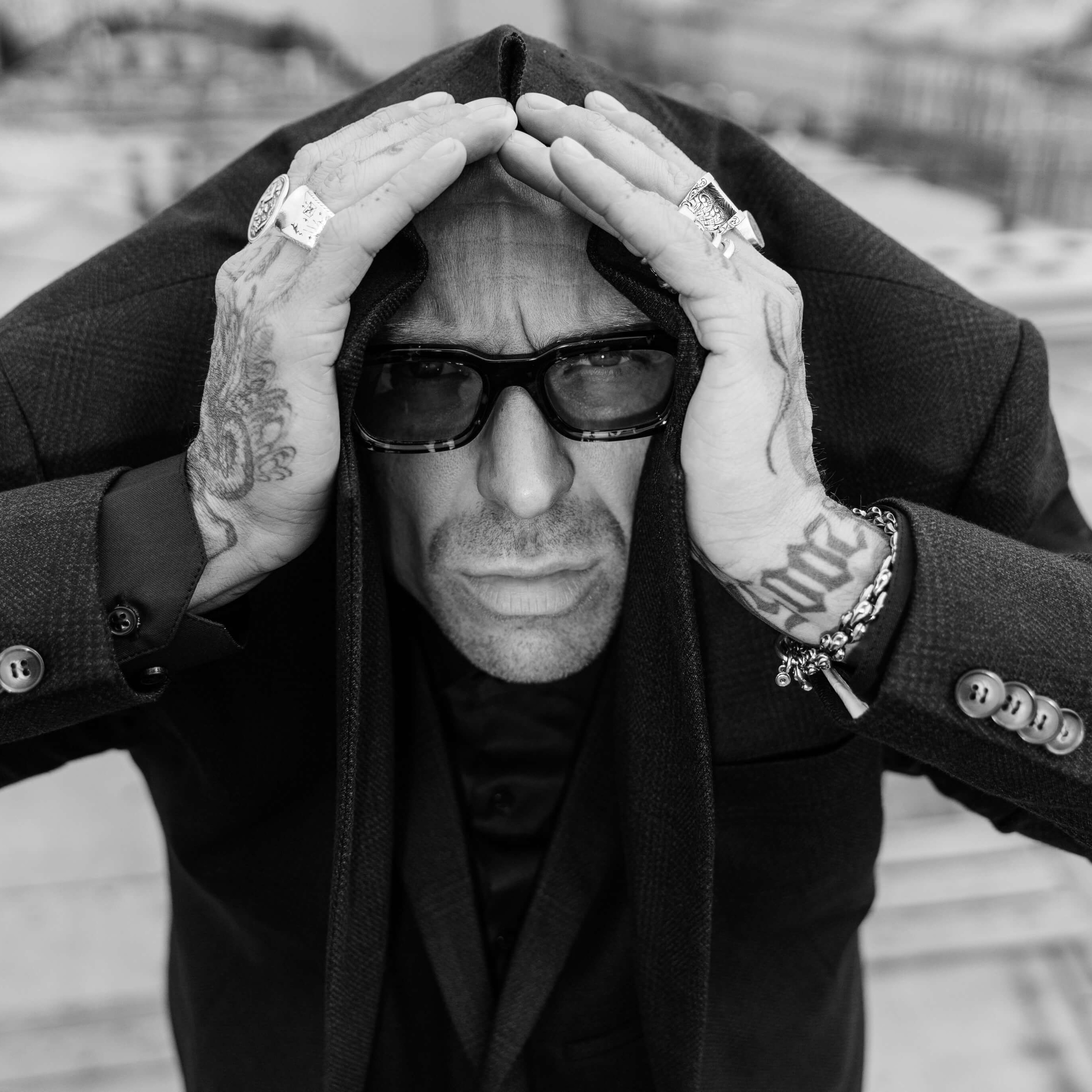 A monochrome image capturing a man in a hooded coat and sunglasses framing his face with his hands, adorned with bold rings and tattoos, conveying a strong, introspective presence against a blurred cityscape.