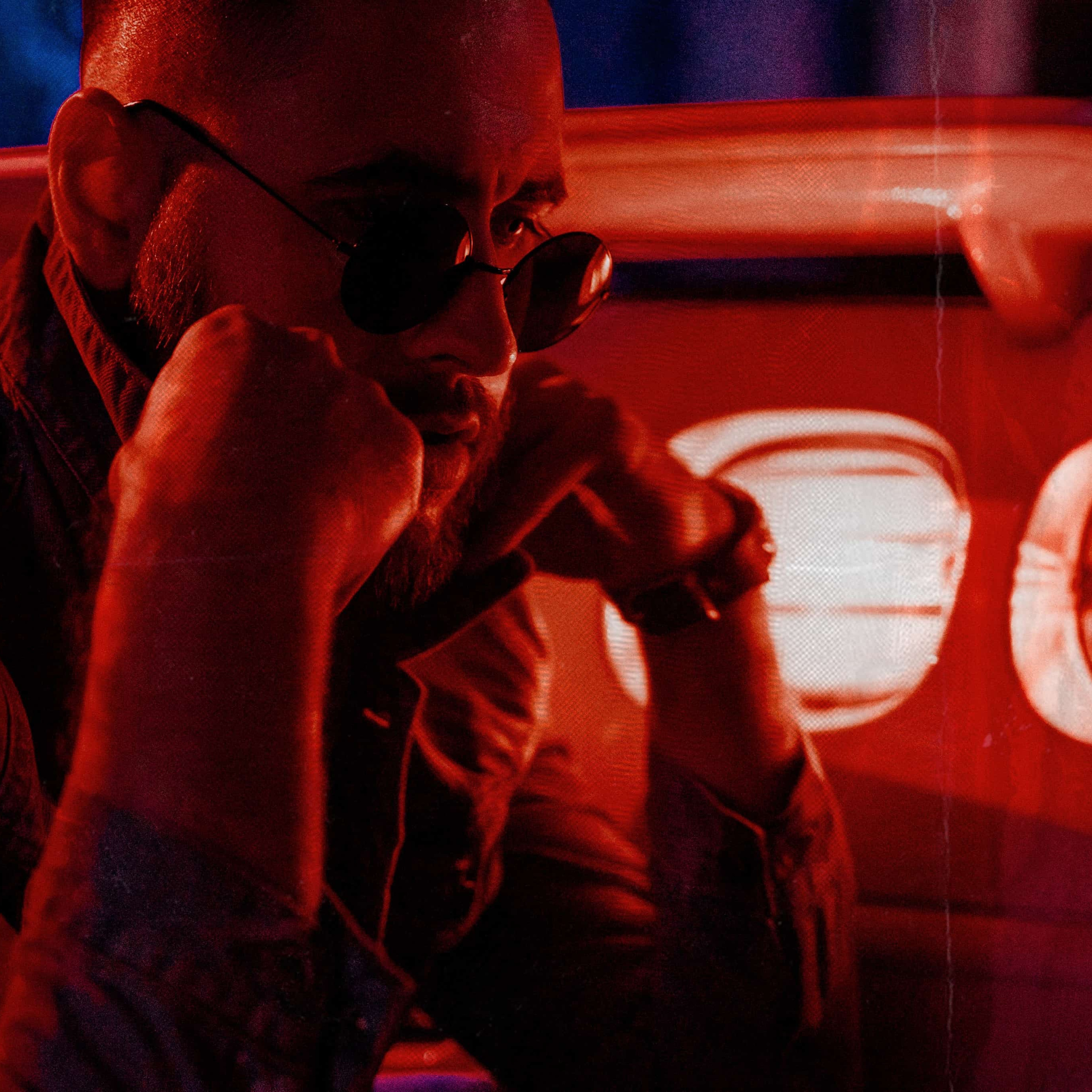 Atmospheric shot of a contemplative man in sunglasses, set against the backdrop of a car's glowing taillights, enveloped in a deep red neon glow