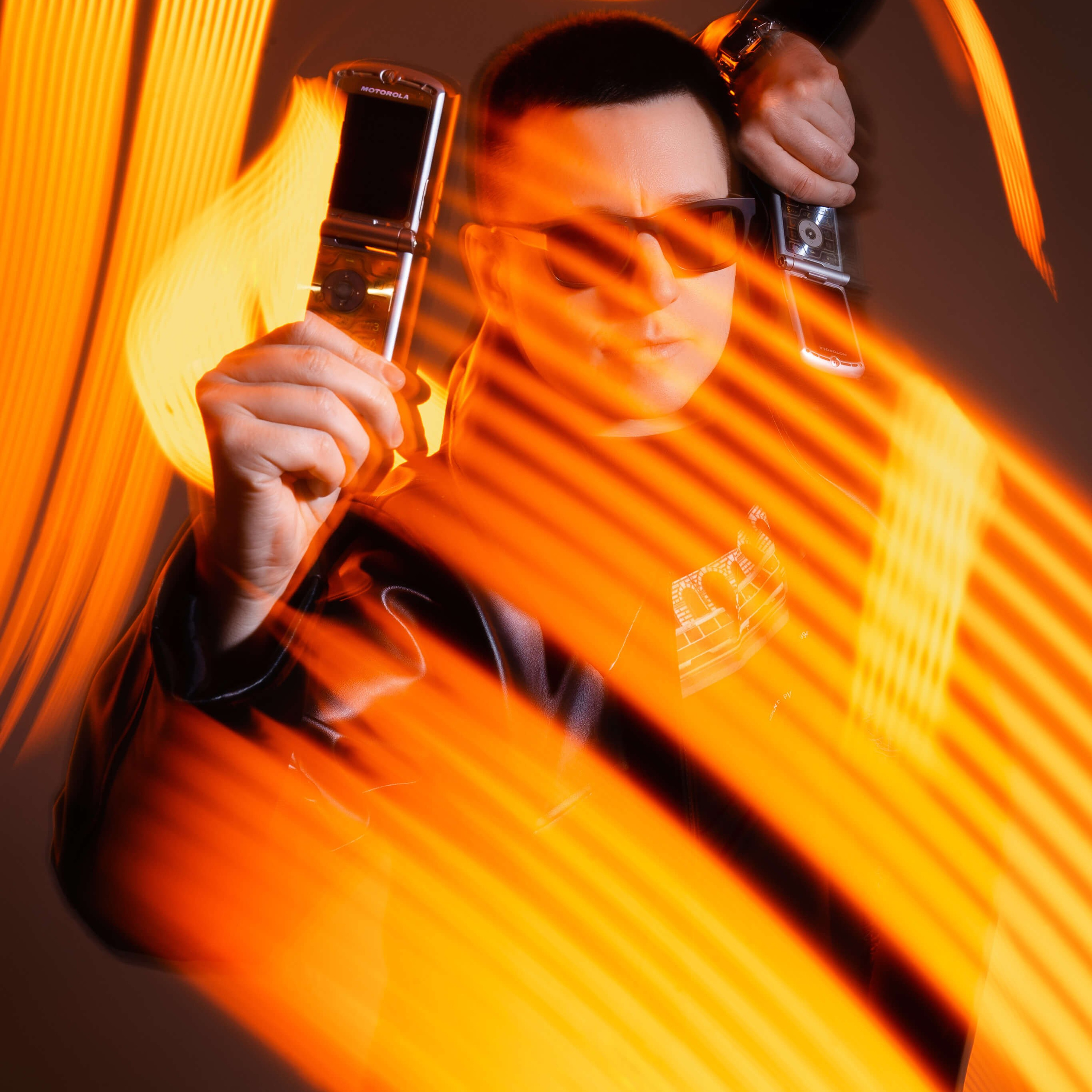 A dynamic image of a person in sunglasses and a black outfit, holding two retro flip phones, with motion blur and orange light streaks conveying a sense of movement.