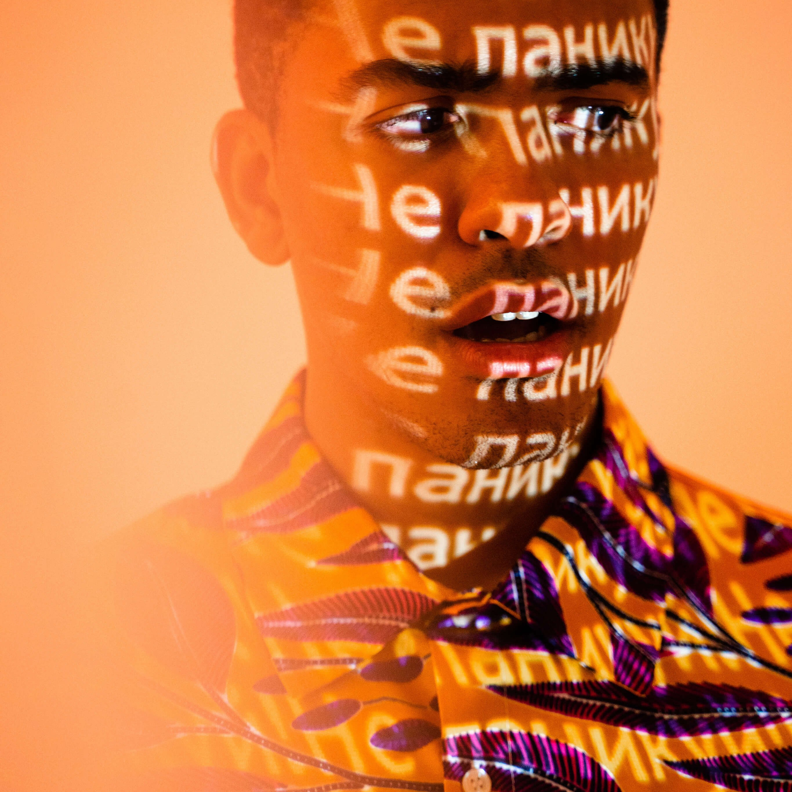 Portrait of a person with text projections casting a pattern over their face and vibrant, patterned shirt, creating a striking interplay of color and typography on the skin, suggestive of a strong personal narrative