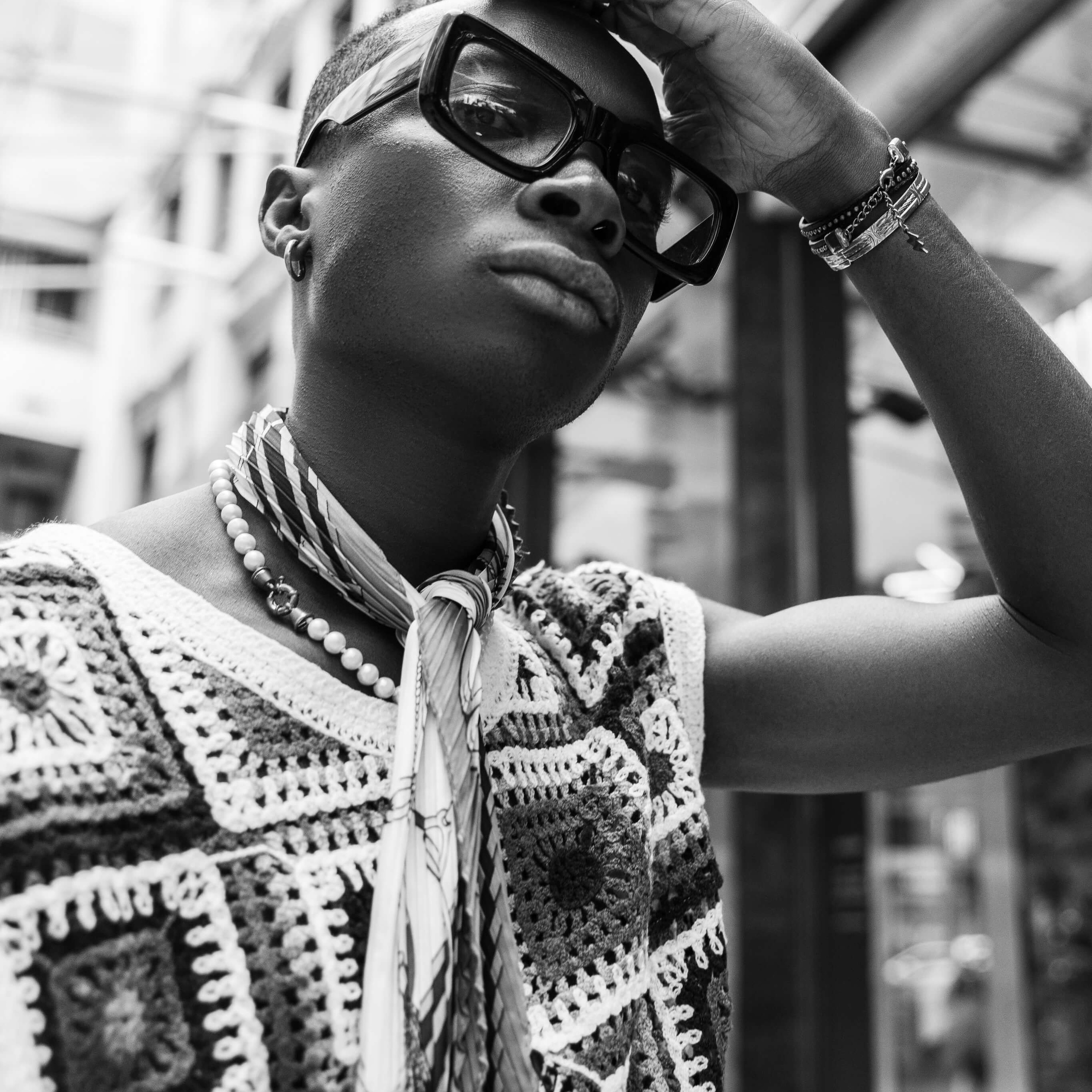 A monochrome portrait of a stylish individual in eyeglasses and a textured crochet garment, shading their eyes and gazing into the distance, with an architectural glass structure providing a modern backdrop