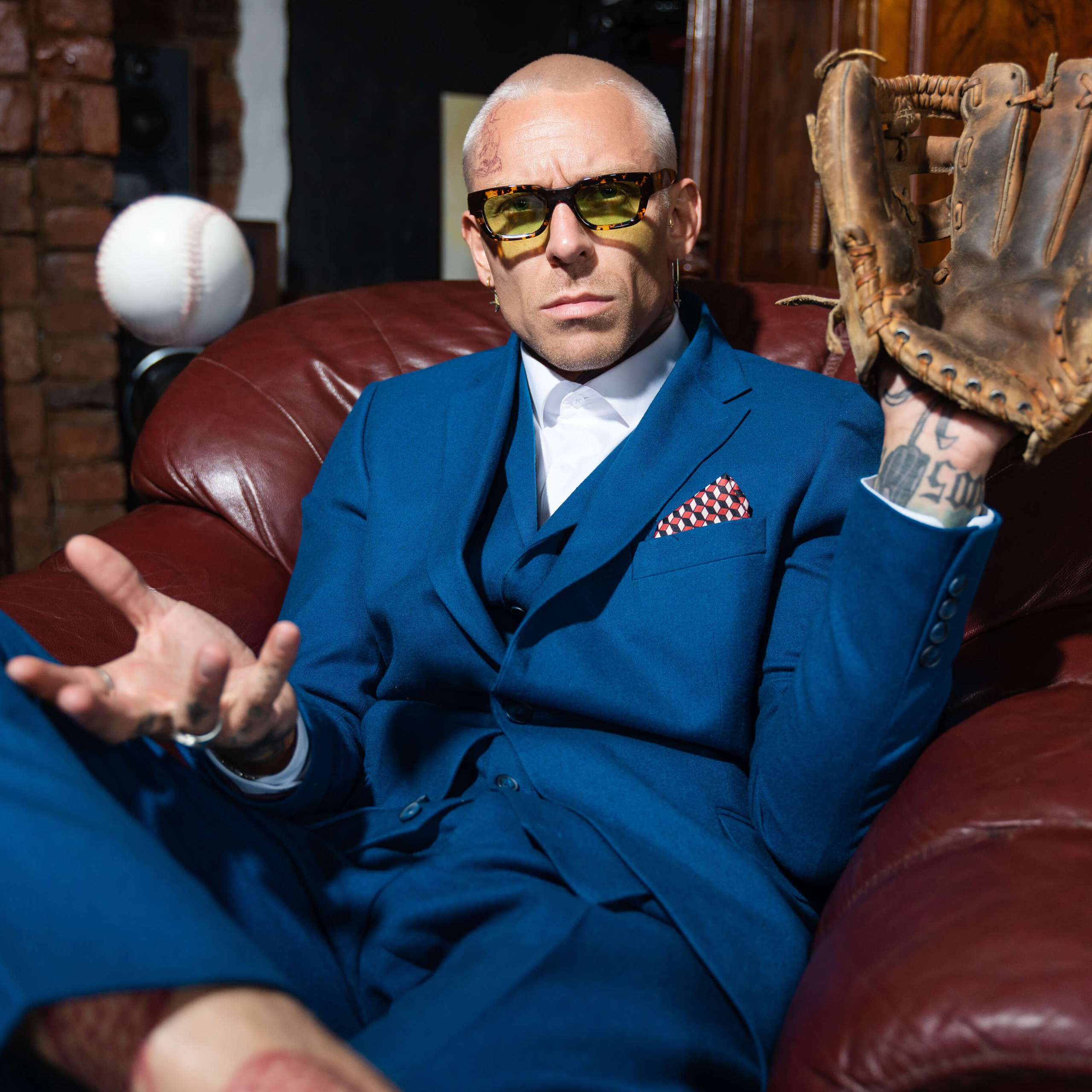 A fashion-forward individual in a sharp blue suit and yellow-tinted glasses, tossing a baseball casually, exuding confidence and a cool demeanor.