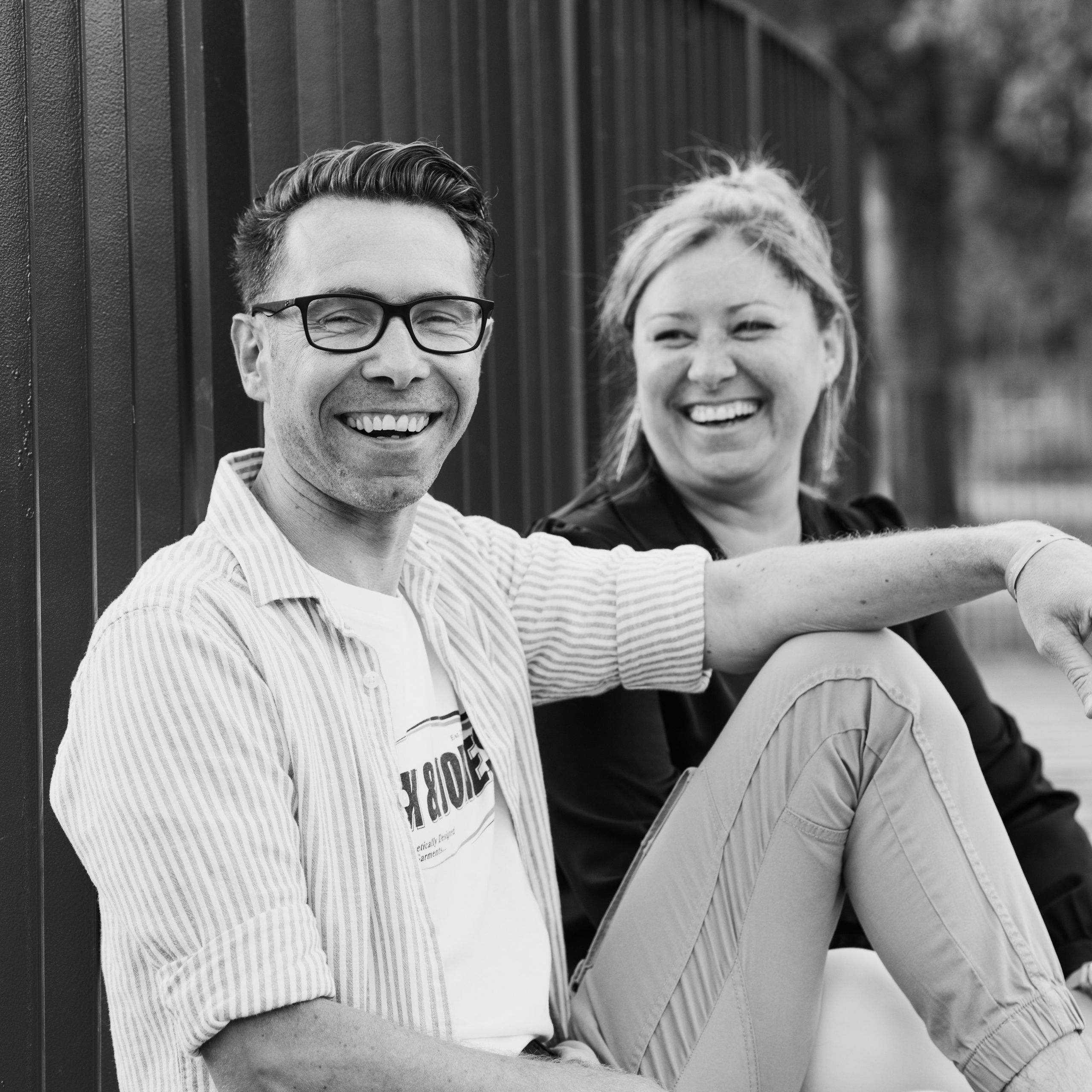 Zwart-wit foto van broer en zus zittend naast elkaar en lachend tijdens spontane familieshoot  - Kapture Fotografie