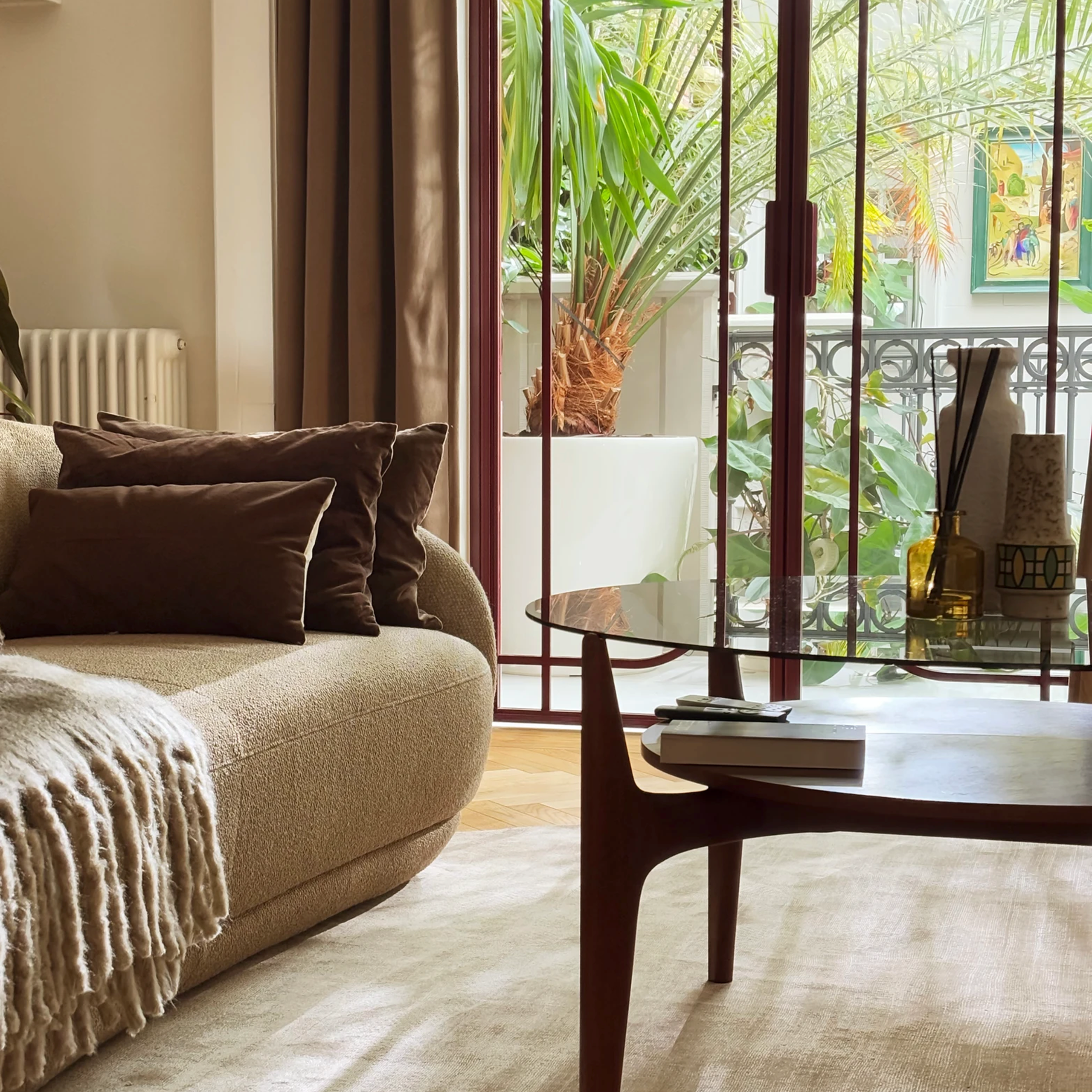 Przytulny salon z beżową sofą, szklanym stolikiem i dużymi oknami z widokiem na tropikalne rośliny.