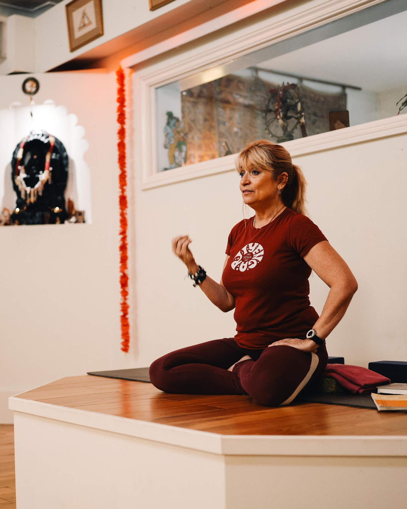 Jayne Orton in Iyengar Yoga Institute of Birmingham. A lady in a red t-shirt is sitting on the platform at the yoga studio.  Yoga brand photoshoot.