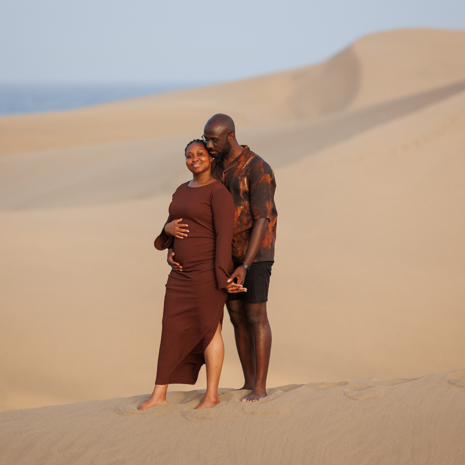 Maspalomas, Grande Canarie. Une femme enceinte vêtue d'une robe marron se tient sur des dunes de sable, souriante, tandis qu'un homme se tient derrière elle et lui tient la main.