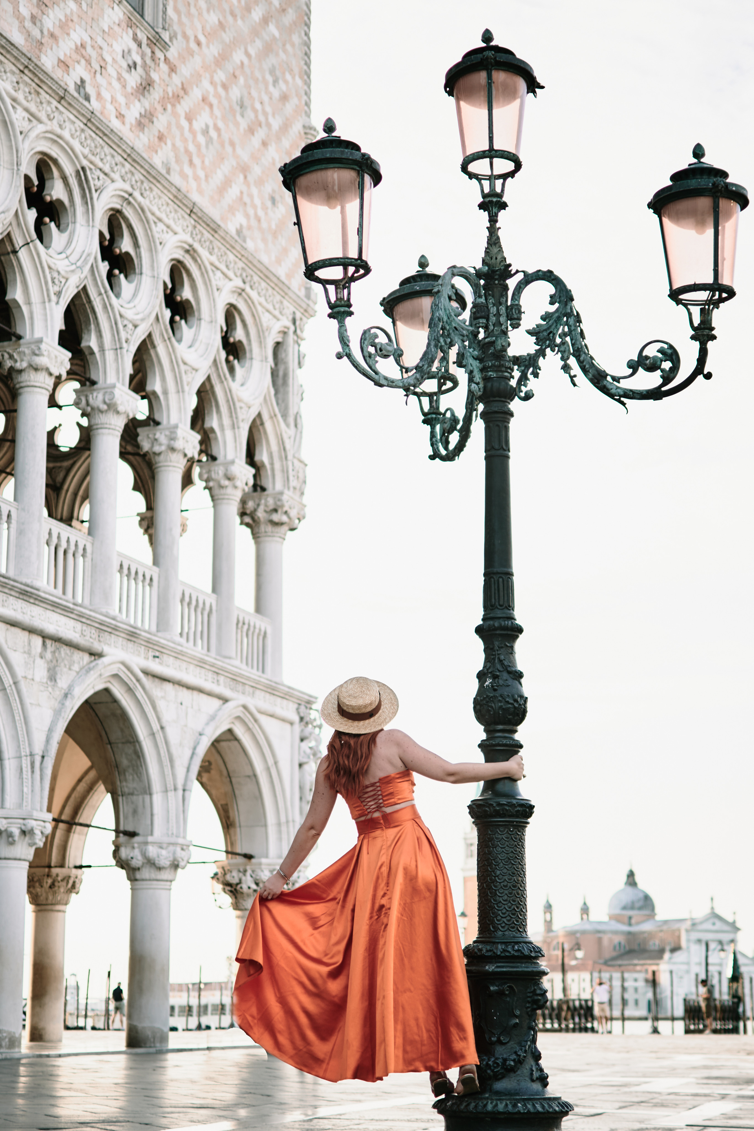 A young woman stands in the heart of San Marco Square in Venice, wearing a flying orange dress that accentuates her beauty. The panoramic view of the Laguna in the background adds to the romantic atmosphere of the scene. The ornate architecture of the surrounding buildings is a testament to the city's rich history and culture. The woman's hair flows gently in the breeze as she gazes out at the stunning view, lost in thought. The vibrant energy of the bustling square is palpable, making for a truly memorable photo shoot.