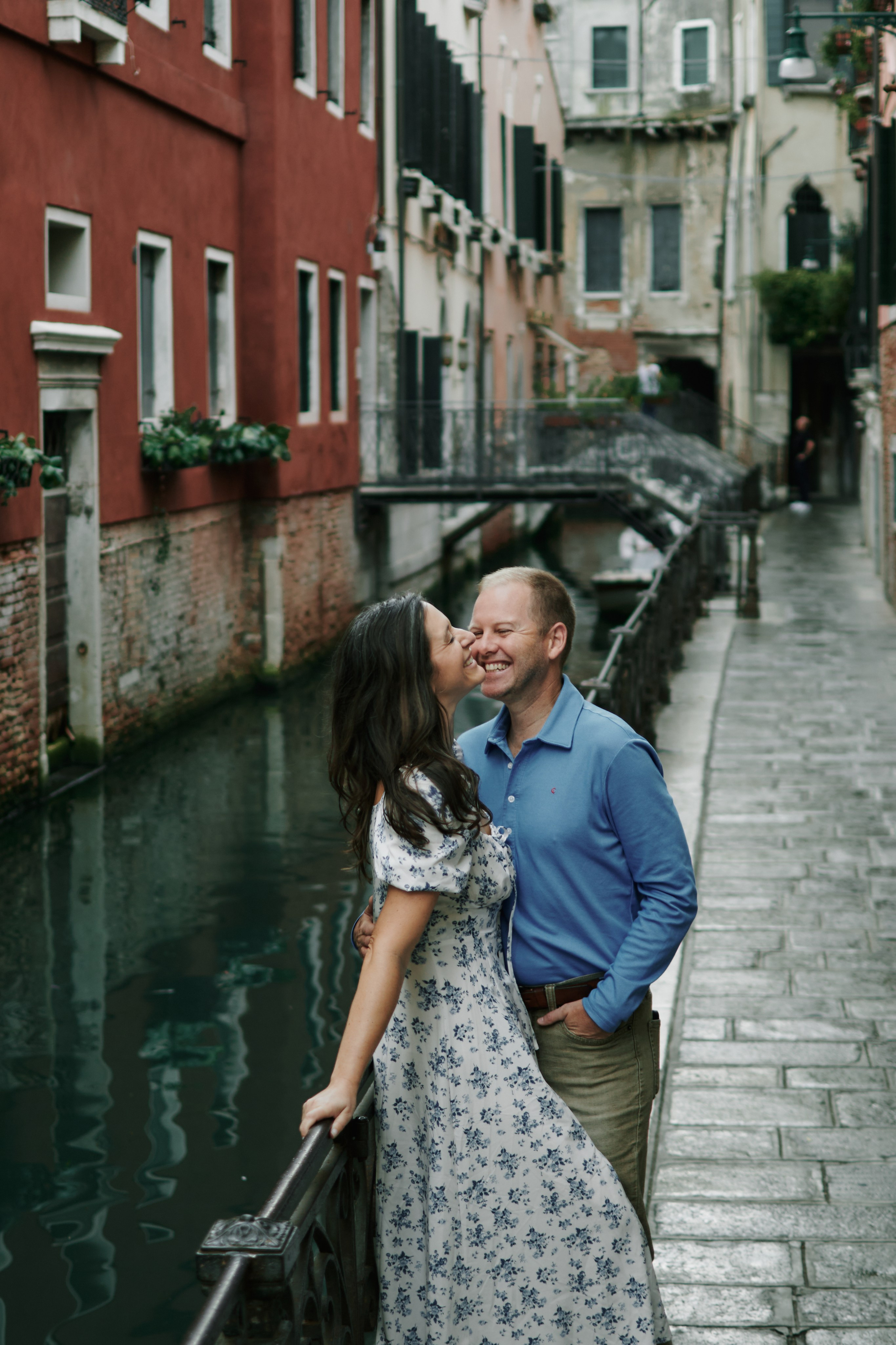 an off-the-beaten-path area  A romantic portrait captures a couple in the midst of a love story shoot in Venice, Italy. They stand close together, gazing out at the canals n the distance. The photographer has expertly framed the shot to showcase both the couple's love and the beauty of Venice's architecture
