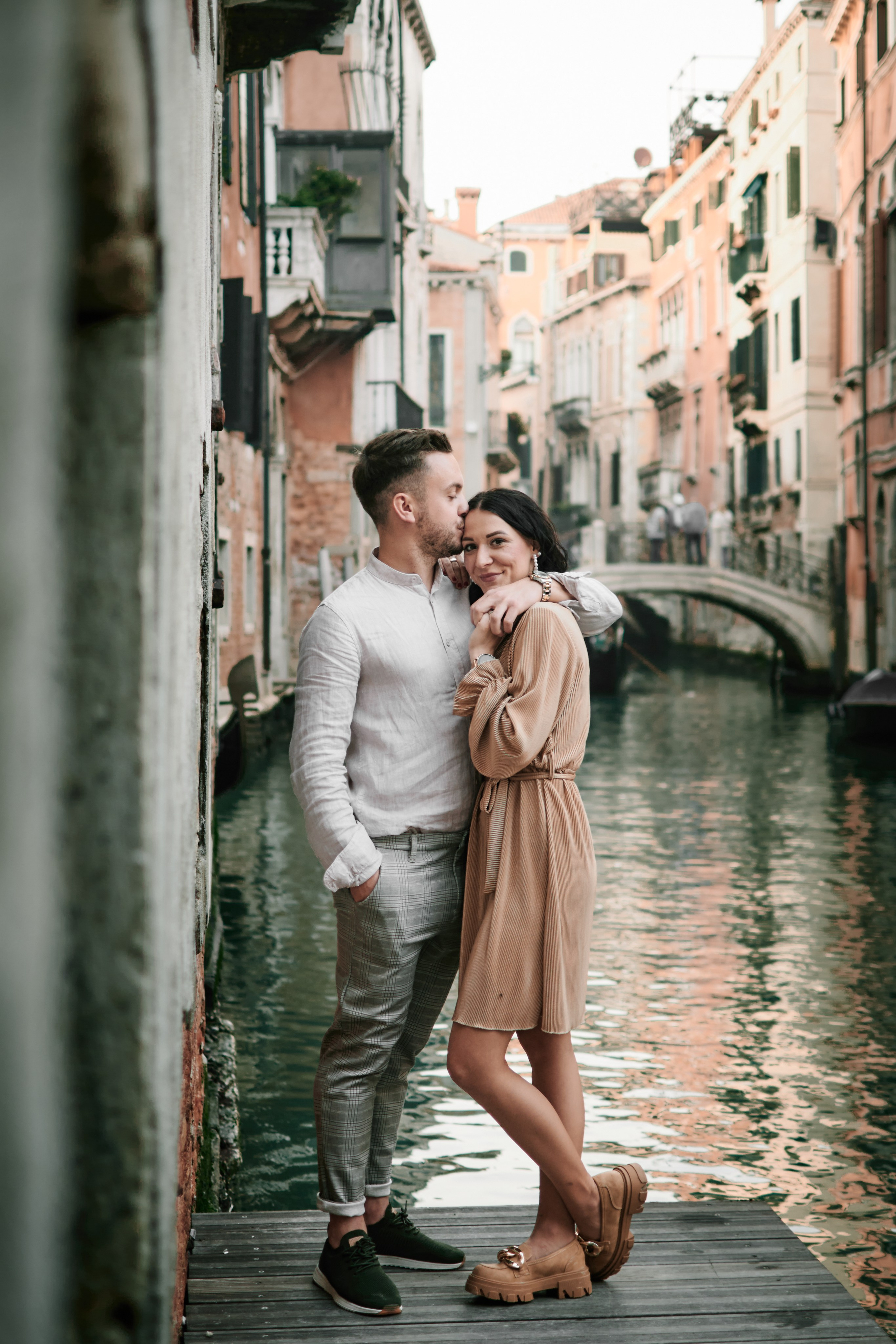 A photographer in Venice, Italy taking a portrait of a person wearing a colorful carnival mask during a photoshoot. The photographer is crouching down with a camera in hand, while the subject stands in front of a canal with the iconic Venetian architecture in the background. The mask features elaborate decorations and intricate designs in shades of red, gold, and blue.