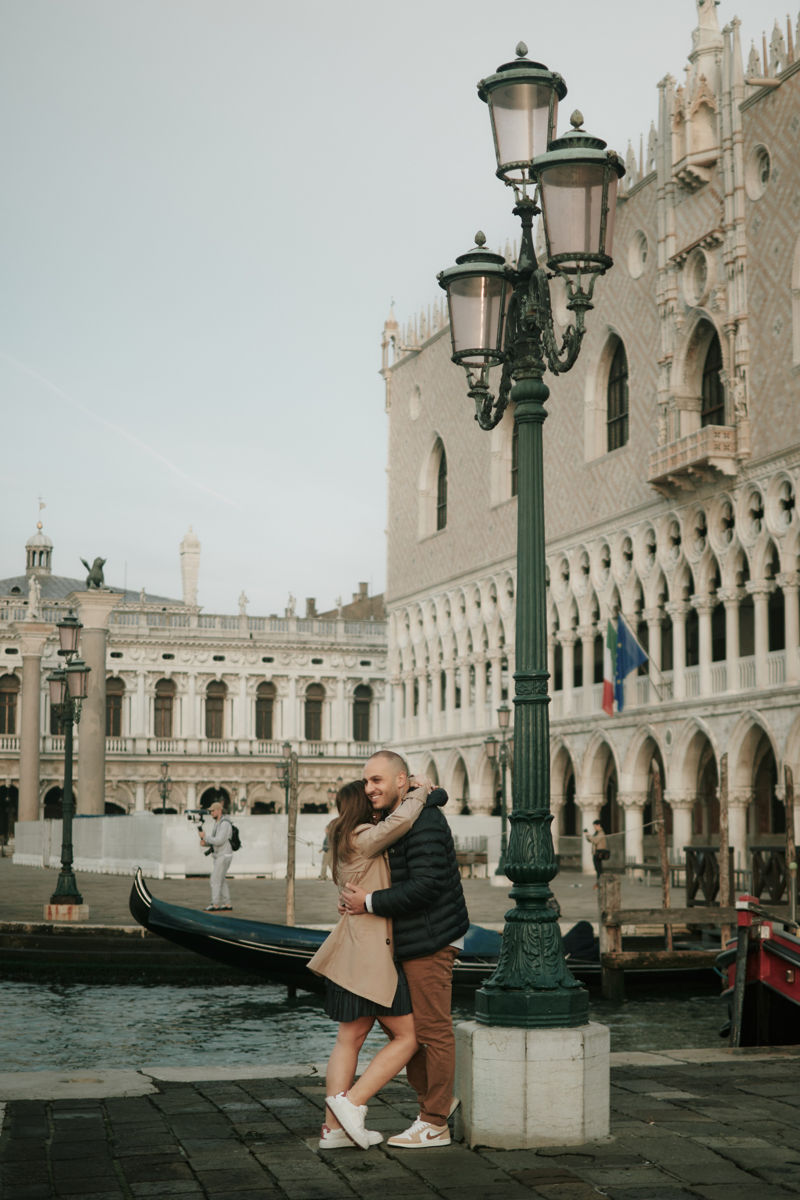 an off-the-beaten-path area. A family of four stands on a small boat on a canal in Venice. The father stands at the back of the boat, holding a long pole, while the mother sits next to him, holding their baby. The older child stands at the front of the boat, gazing at the beautiful Venetian architecture in the background. The family is surrounded by calm waters and colorful buildings, creating a serene atmosphere. The photo captures the essence of a peaceful family moment amidst the picturesque backdrop of Venice