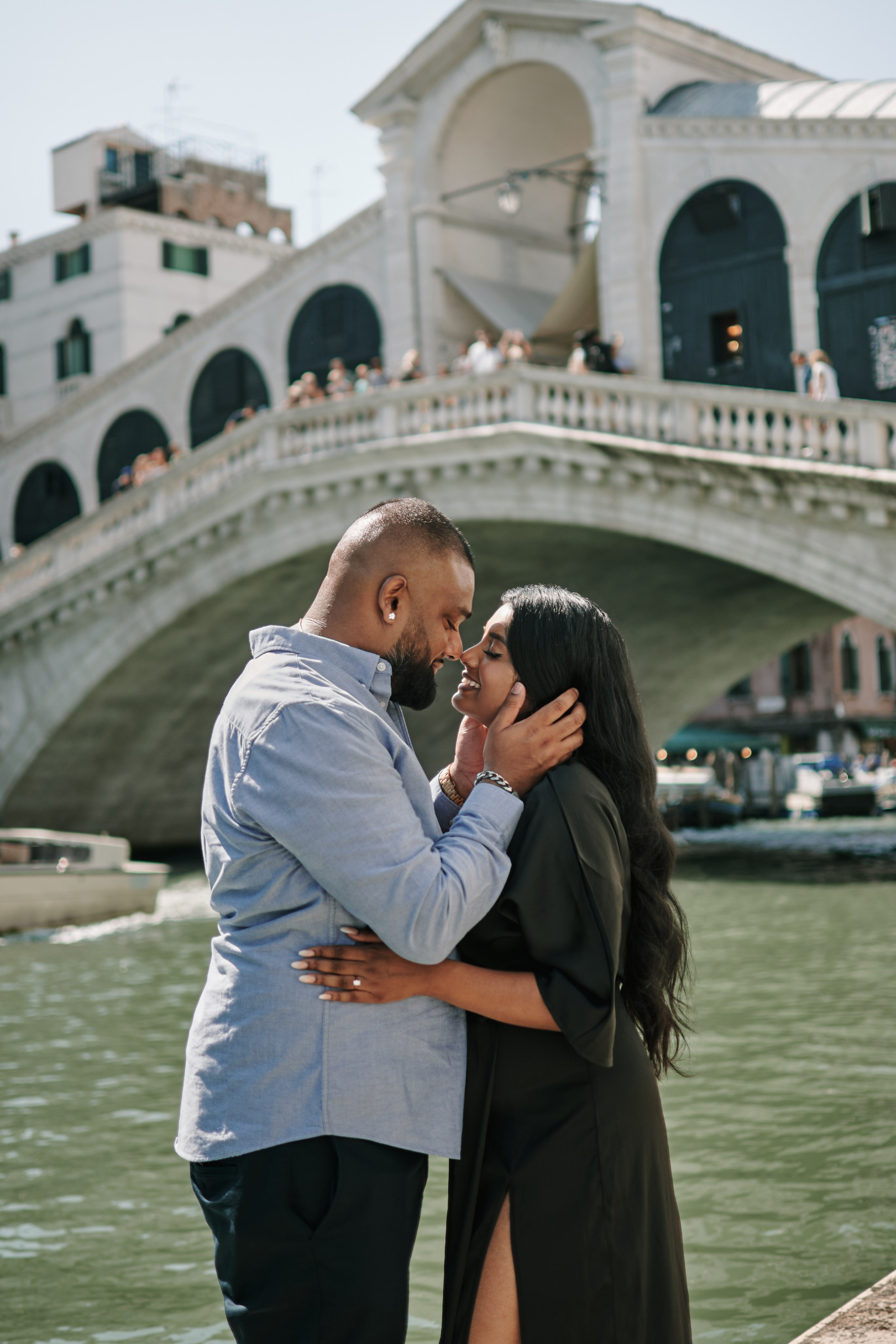 A young woman stands tall in front of the grand canal near Rialto Bridge in Venice, Italy. The woman's long hair flows freely in the gentle breeze as she gazes gracefully into the distance. The towering columns, adorned with intricate carvings, frame the woman beautifully, creating a striking contrast between the ancient architecture and the woman's youthful appearance. The warm, golden light of the setting sun bathes the scene, casting a soft glow on the woman's face and the majestic columns behind her.