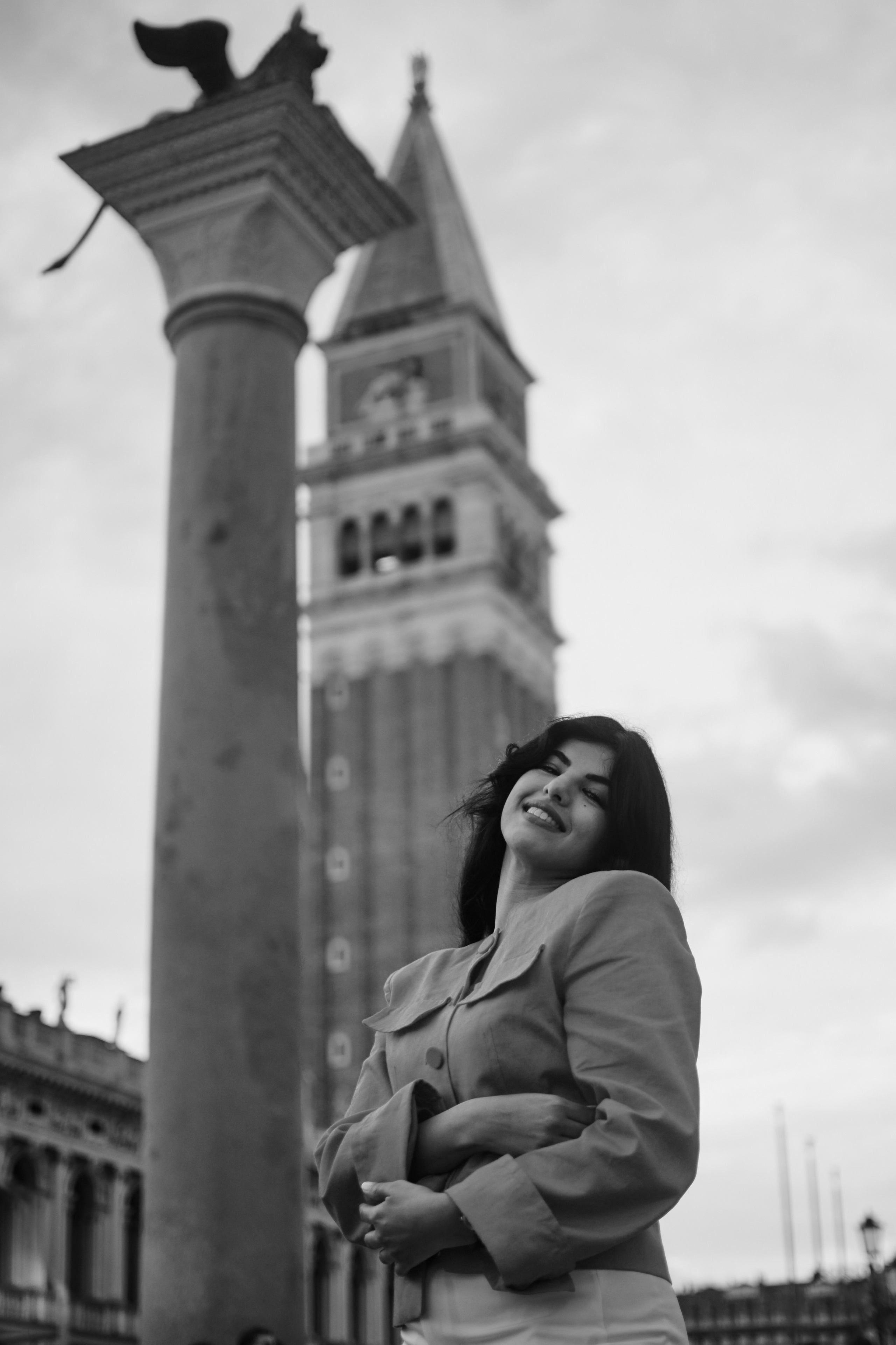 This portrait photo features a young woman on the San Marco square in Venice, with the towering St. Mark's Basilica rising up behind her. The woman looks off into the distance, lost in thought. The blue sky and warm sun give the photo a serene and peaceful feel. Gondolier woman, venice pier