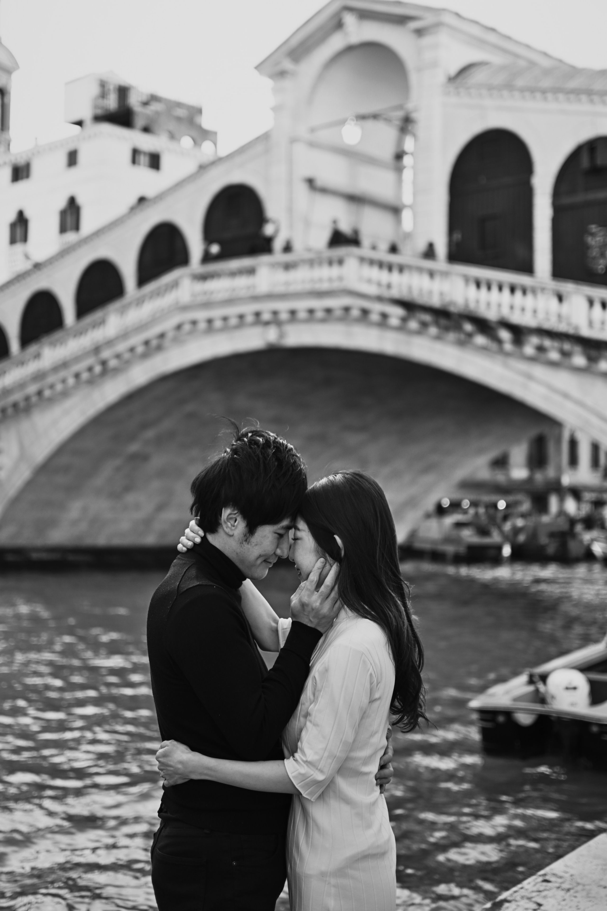 In this beautiful portrait photo, a couple stands hand-in-hand, gazing out at the breathtaking panorama of the lagoon and Rialto Bridge. The intricate details of the surrounding architecture and the warm, golden light of the setting sun create an atmosphere of beauty, love, and passion. Black and white photography in Italy