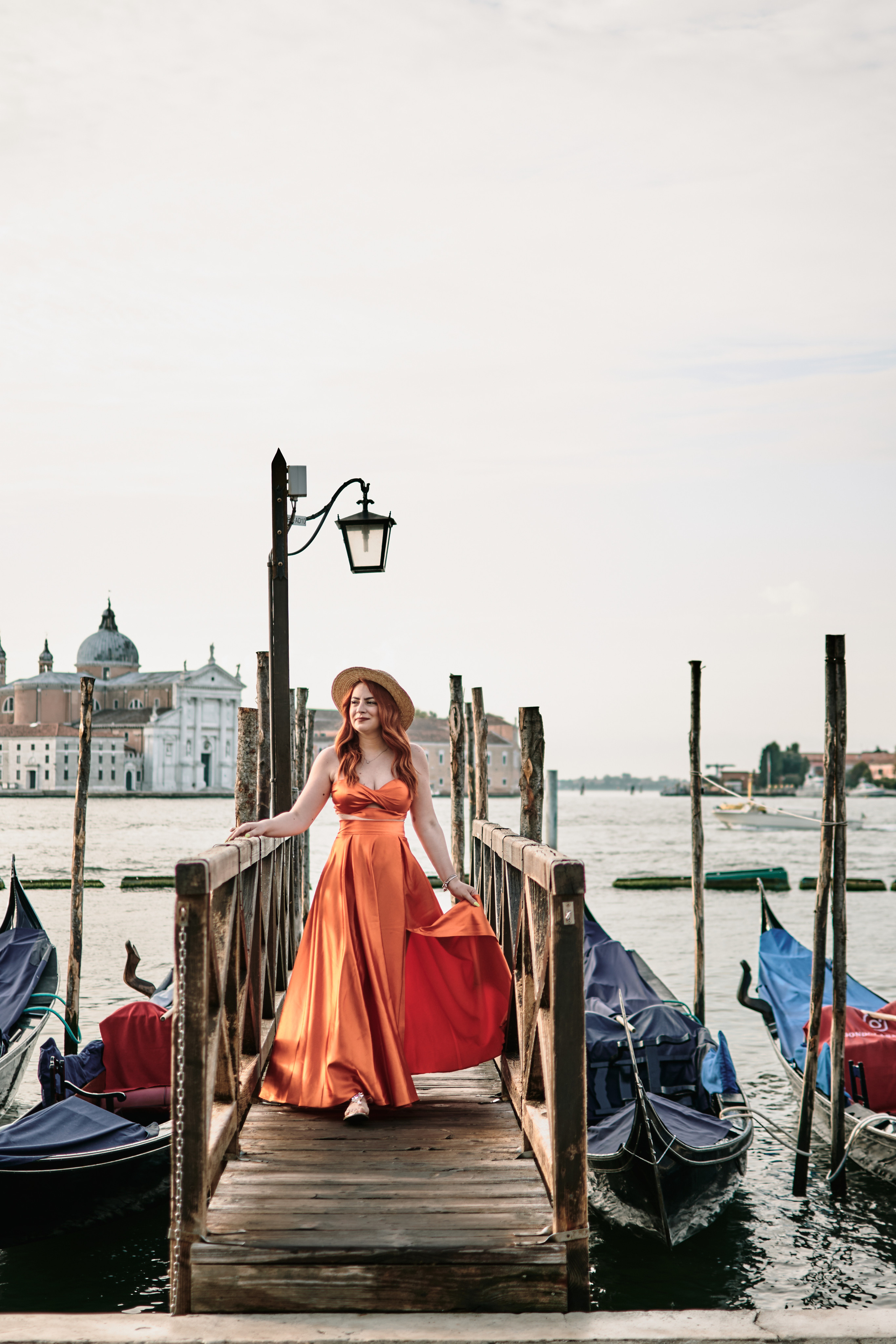 A beautiful young woman, stands in front of the iconic Venetian backdrop of San Marco square. She wears a flowing  dress that accentuates her natural beauty. The soft light of the sun illuminates her face, and her serene expression captures the joy . The tranquil waters of the canal behind her add to the peaceful atmosphere of this stunning portrait
