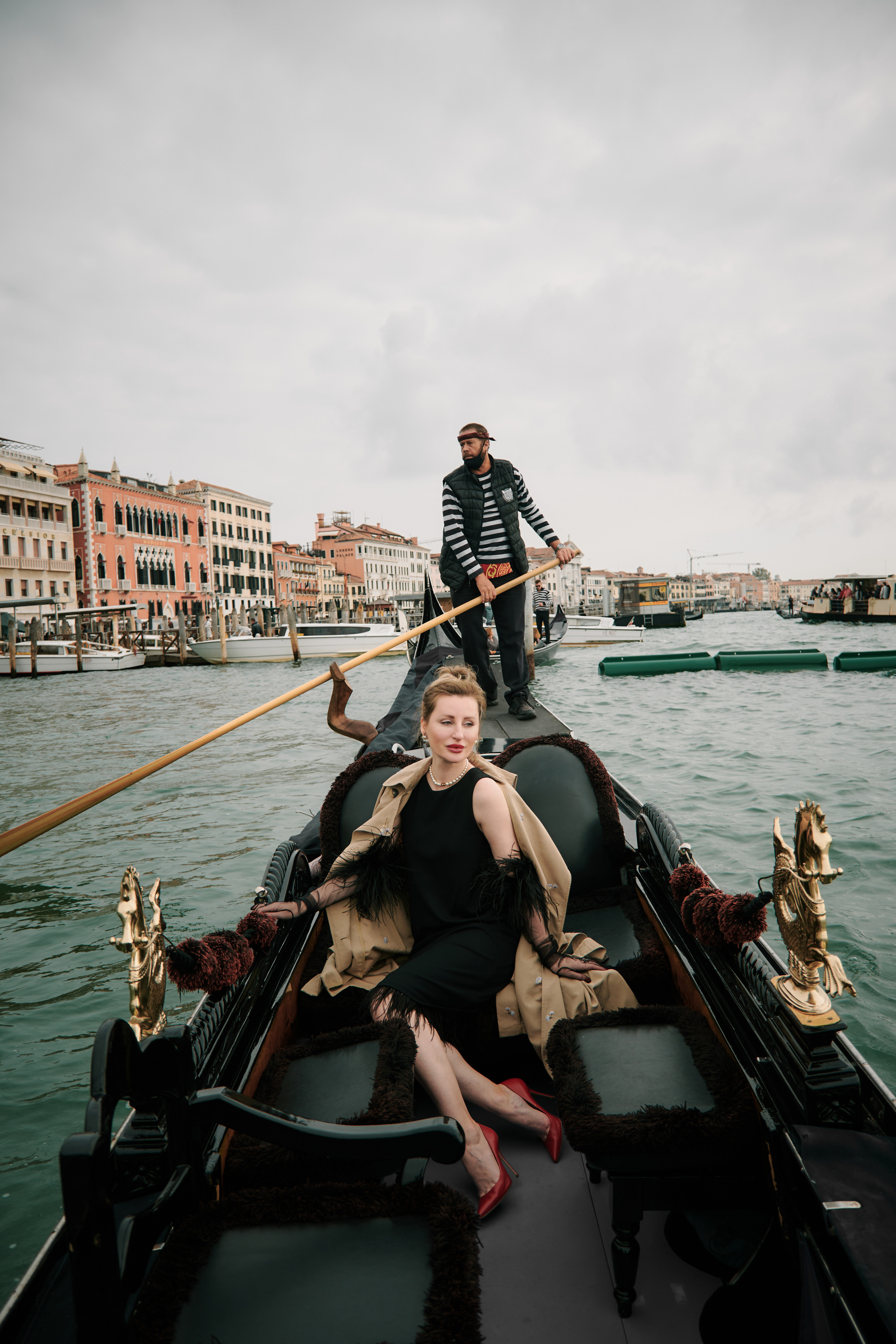A young woman in a black dress sits on a gondola as it glides through the narrow streets of Venice. She gazes off into the distance, lost in thought, as the sunlight dances on the water around her. The picturesque buildings of the city rise up on either side, their pastel colors and ornate details adding to the romantic atmosphere. The woman's elegant dress and captivating presence seem perfectly at home in this enchanting setting, creating a sense of mystery and allure that draws the viewer in