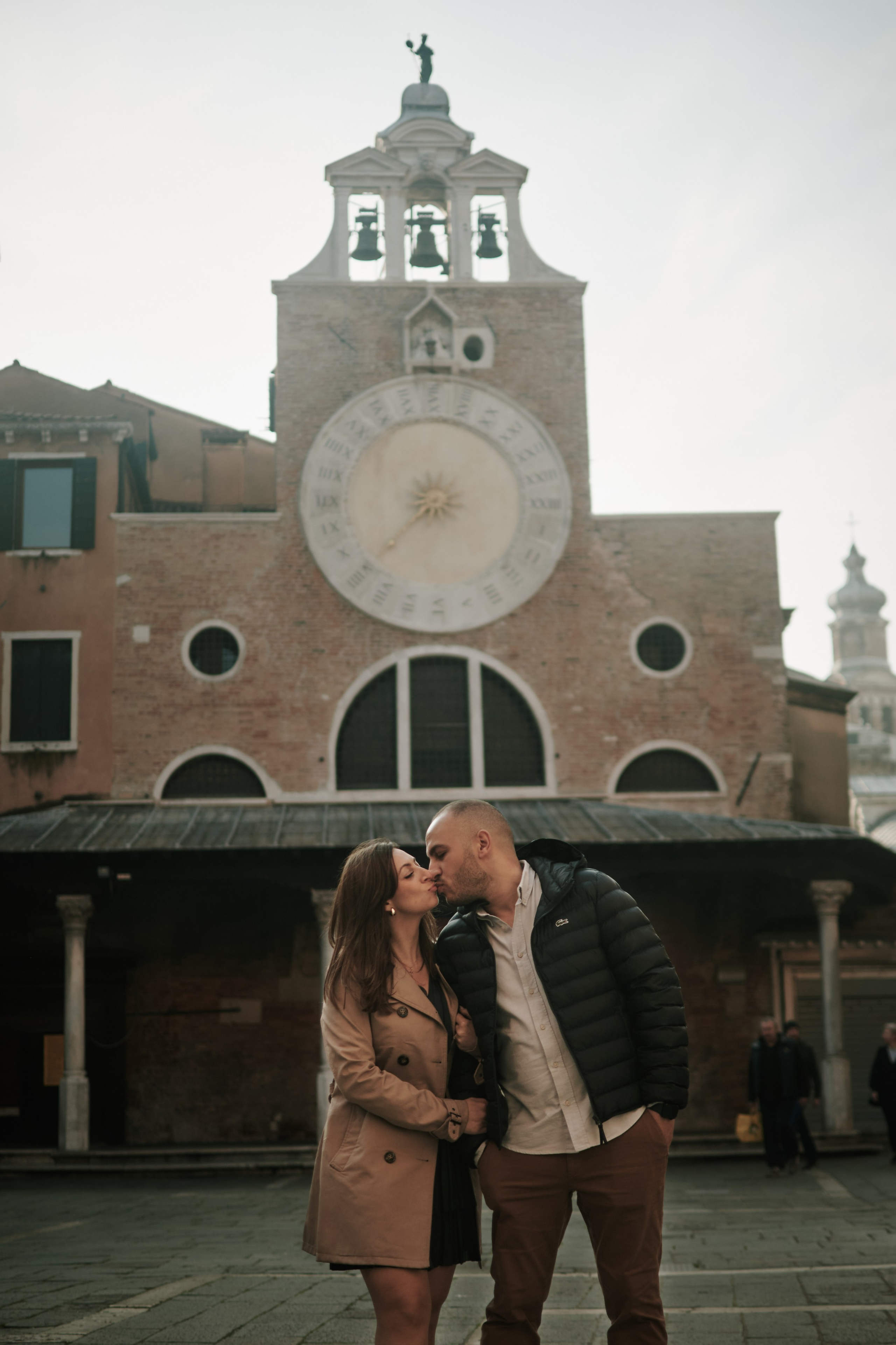 Portrait of a young woman in a black dress standing on Rialto bridge, Venice, with a breathtaking panoramic view of the Laguna in the background. The woman has long brown hair styled in loose waves, and she is looking dreamily towards the view. The square is bustling with people, and the stunning architecture of the surrounding buildings adds to the romantic atmosphere of the location