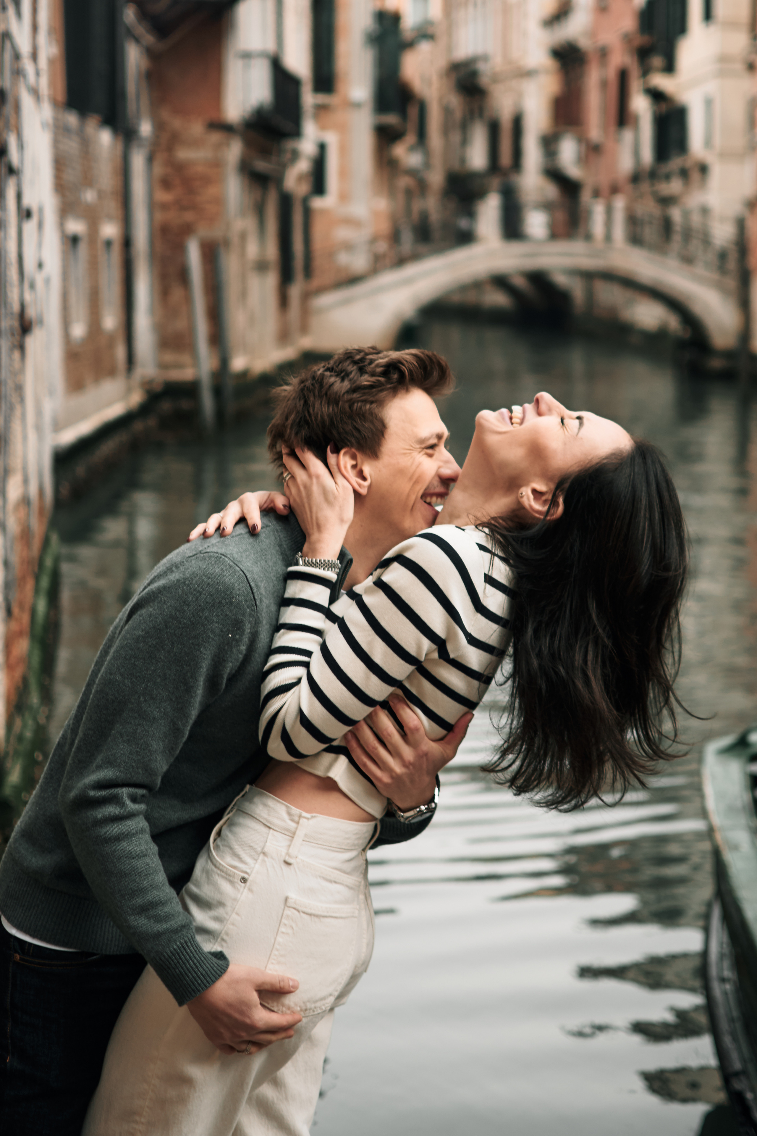 A portrait of a couple in love against the backdrop of Venice's stunning architecture. A happy family  is standing on a bridge in Venice, Italy, with the beautiful Canal in the background. The lovers are holding hands and smiling at each other. The warm sunlight is reflecting off the water, creating a serene and peaceful atmosphere. The architecture of the surrounding buildings is typical of Venice, with pastel-colored walls, arched windows, and red-tiled roofs.