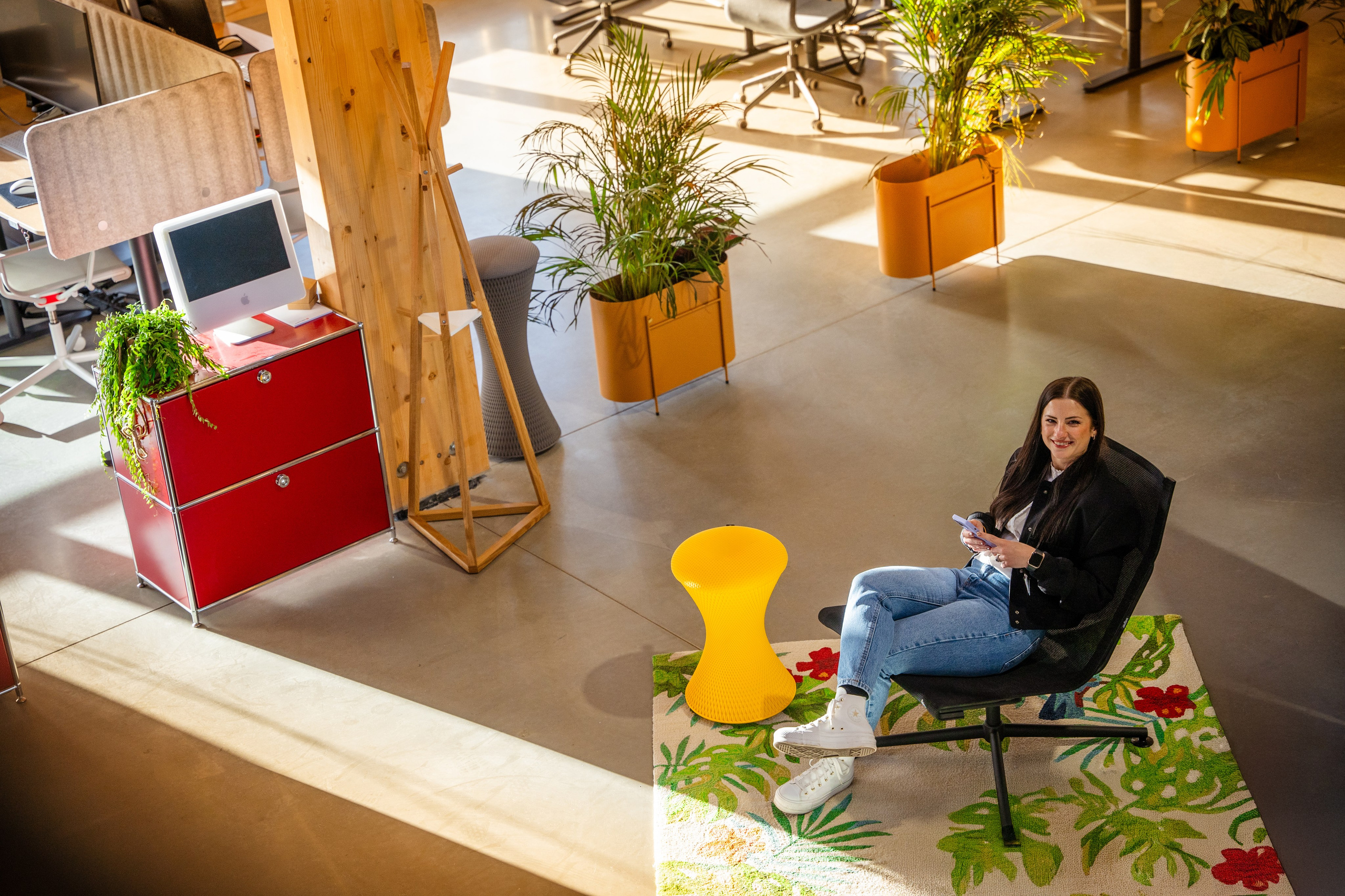 Frau sitzt entspannt auf einem Stuhl in einem modernen, hellen Büro mit Pflanzen und farbenfrohen Möbeln; ein gelber Hocker steht vor ihr, und im Hintergrund sind ein rotes Schrankmodul, ein Computer und weitere grüne Pflanzen zu sehen