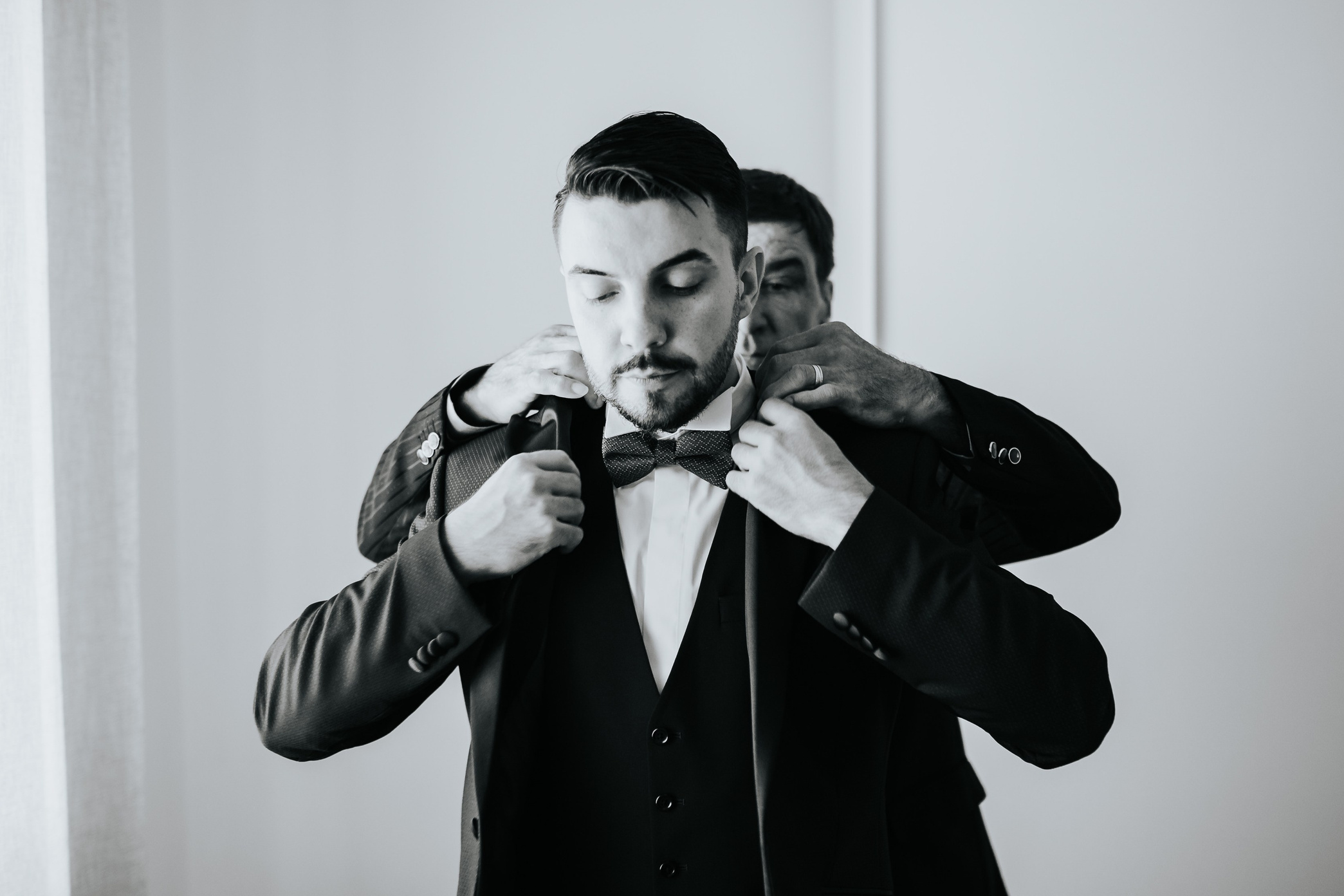Immagine monocromatica catturata da un fotografo matrimonio che ritrae lo sposo nell'intimo momento della preparazione, sistemando il suo papillon davanti a uno specchio. Il dettaglio in bianco e nero accentua l'eleganza del vestito e la serietà dell'evento imminente, offrendo uno sguardo sulle tradizioni e le emozioni del giorno del matrimonio.