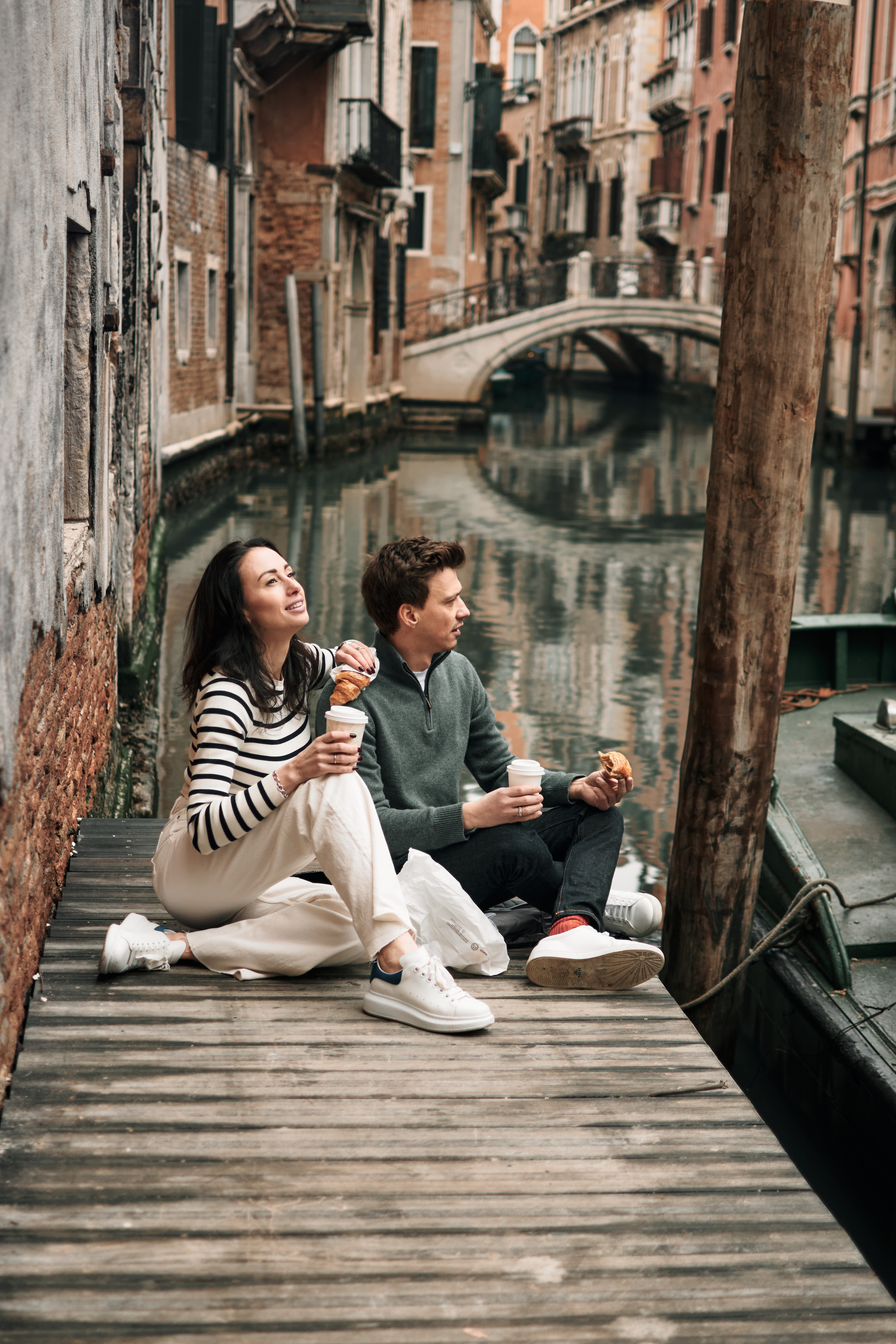A family poses in front of a Venetian bridge, capturing their memorable vacation in Venice. Couple eats croissants in Venice, Italy. A loving couple posing in a narrow alleyway in Venice, Italy, with colorful buildings and street lamps in the background.