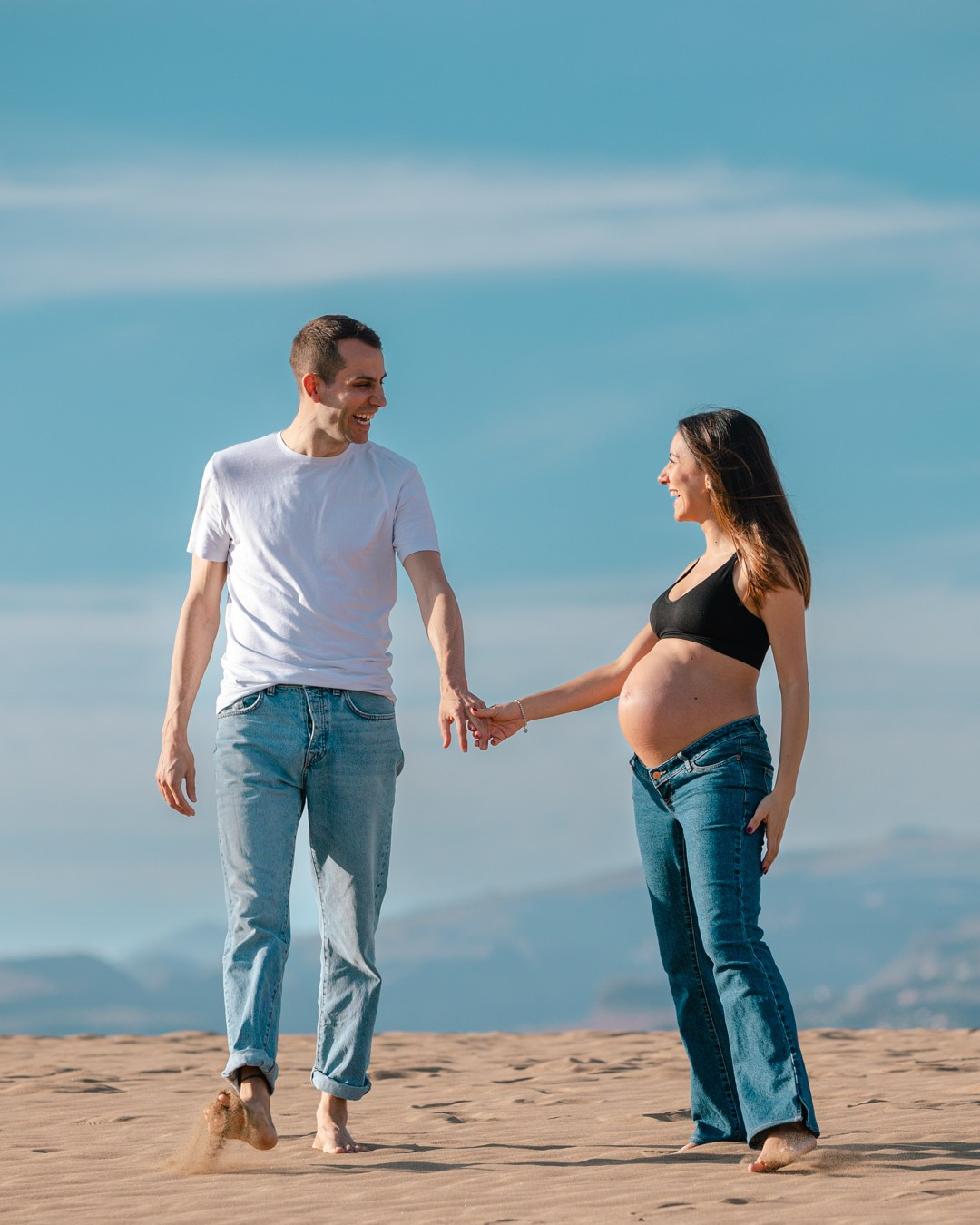 A pregnant couple holding hands in the sand.
