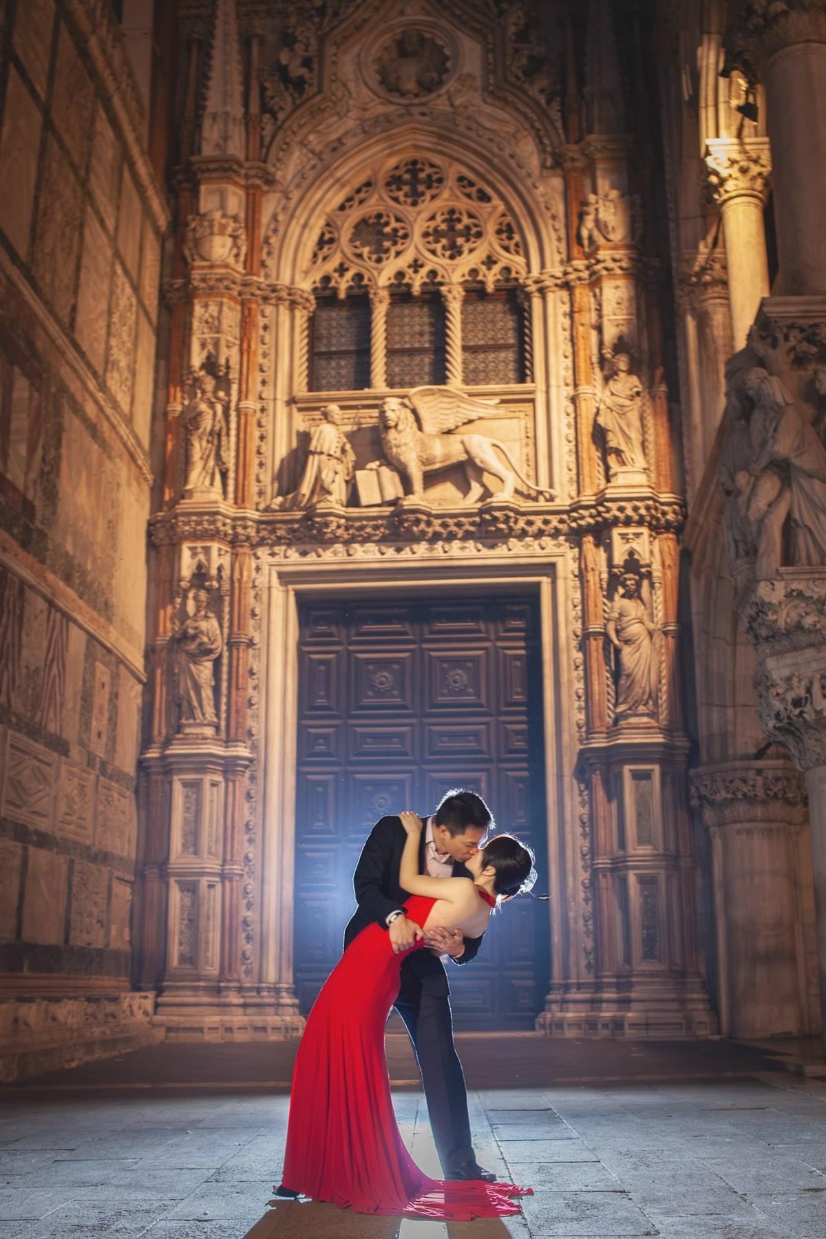 A woman wearing a red dress is dipped and kissed by her Thai fiancee near the historic doors of the Doge's Palace in this nighttime portrait where a rim flash was used.