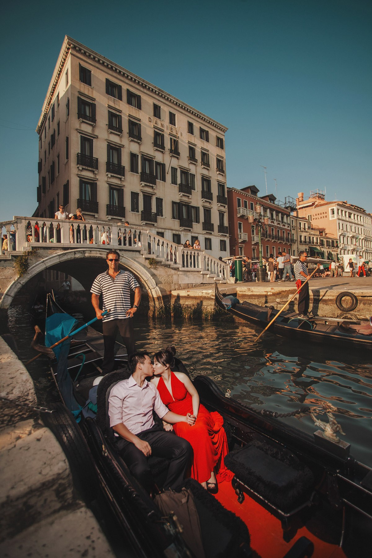 A smiling Thai woman wearing a sexy red evening dress awaits the start of her gondola ride with her fiancee near the Hotel Danieli in Venice during their Love Story Adventures.