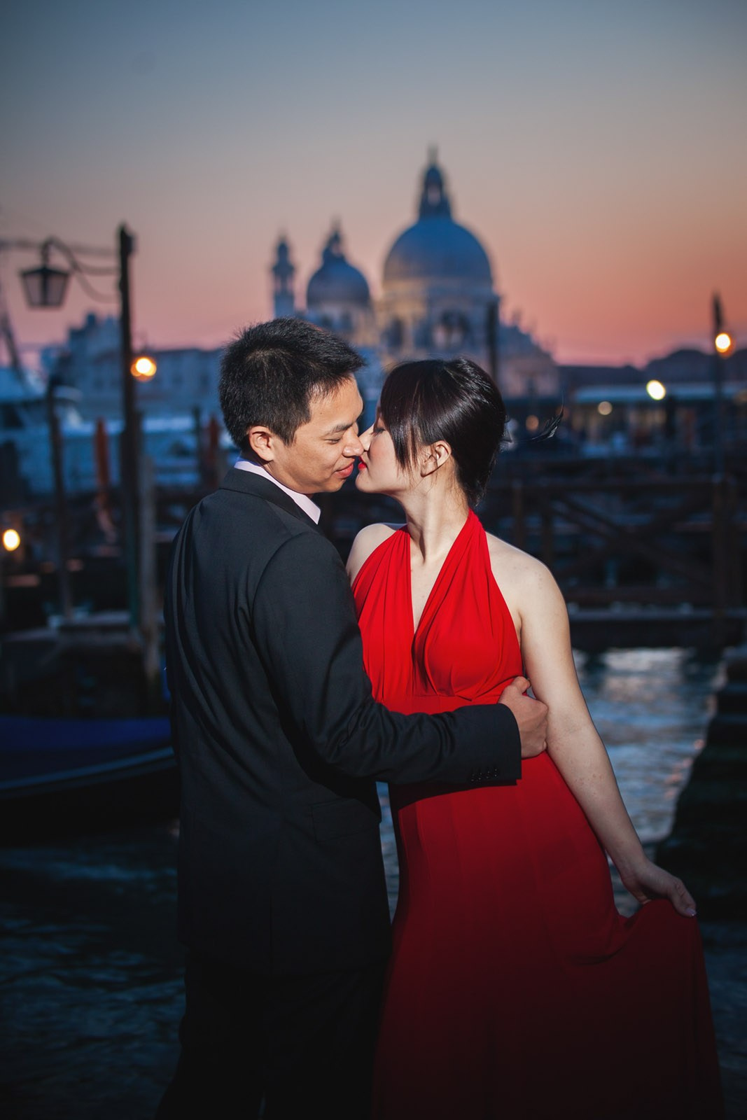 A Thai woman wearing a sexy, skin tight red dress is caressed by her fiancee as they stand near the gondola as the sun sets behind them in Venice.