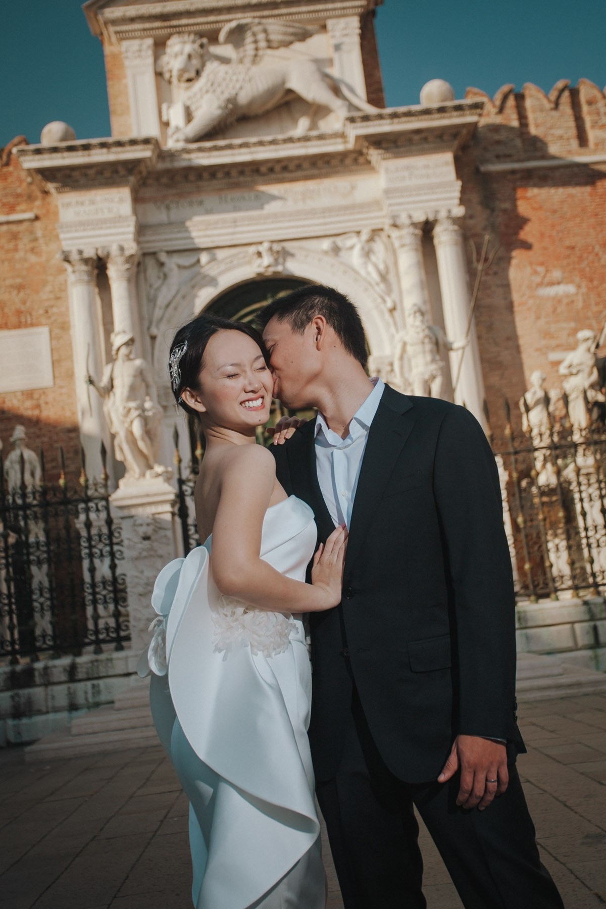 A smiling Thai bride wearing a traditional white wedding dress is kissed by her fiancee in front of the historic Arsenale fortress in historic Venice during their Love Story photo session.