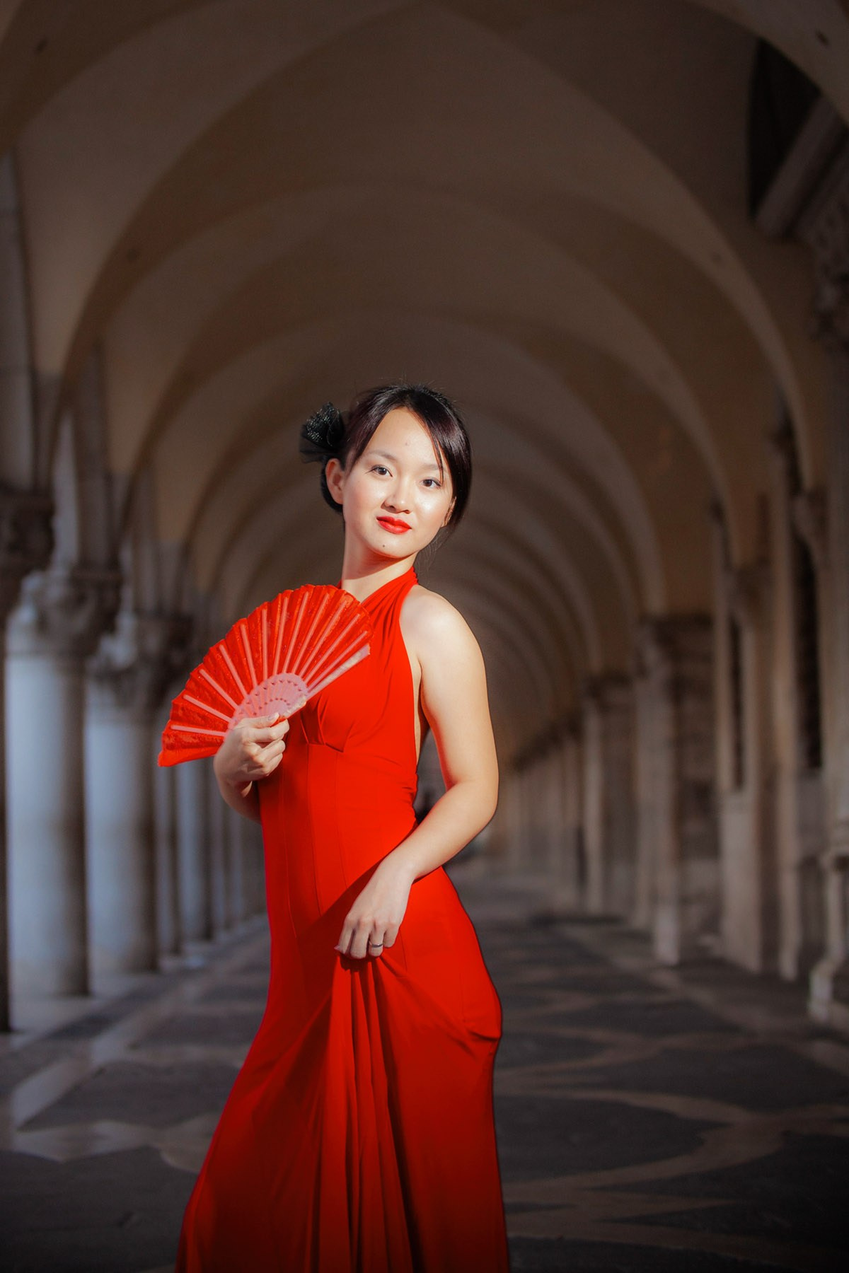 A beautiful Thai bride holding a red fan poses for a portrait under the arches of the historic Doge's Palace as she wears a sexy red dress. 