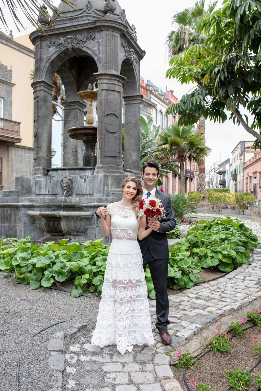 A bride and groom are posing in front of a fountain.