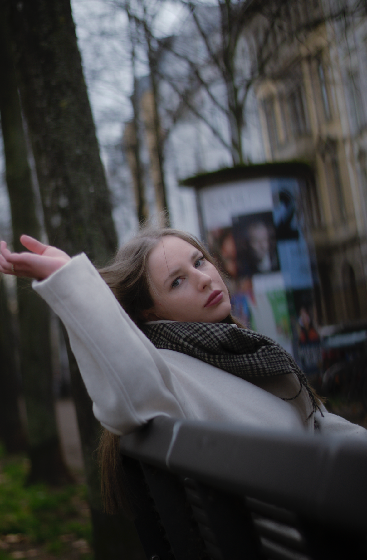 Porträt einer jungen Frau mit dunkelblonden Haaren, blauen Augen und hellbeigem Mantel sitzend auf einer Parkbank