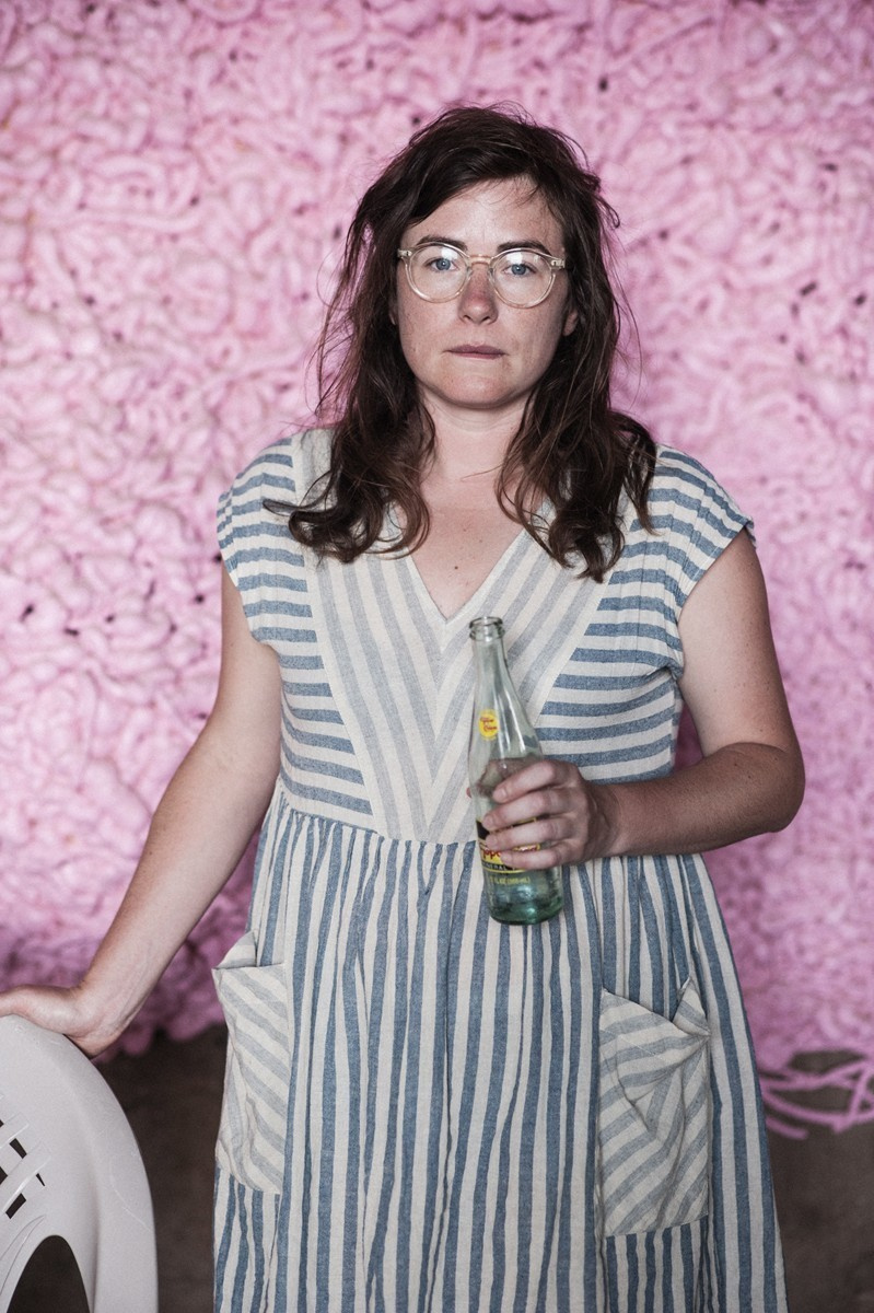 A female on a pink wall wearing a striped board with coke in her hand.