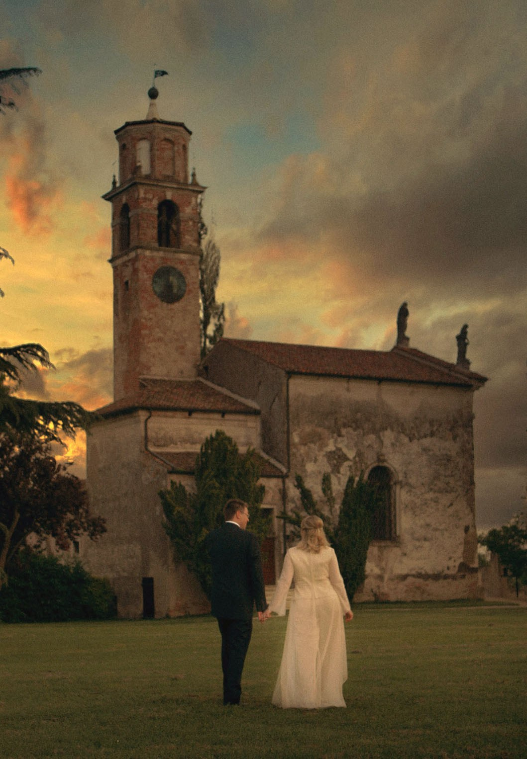 Bride and groom walk across the historic landscape where a 13th-century church stands as the sky flares above.