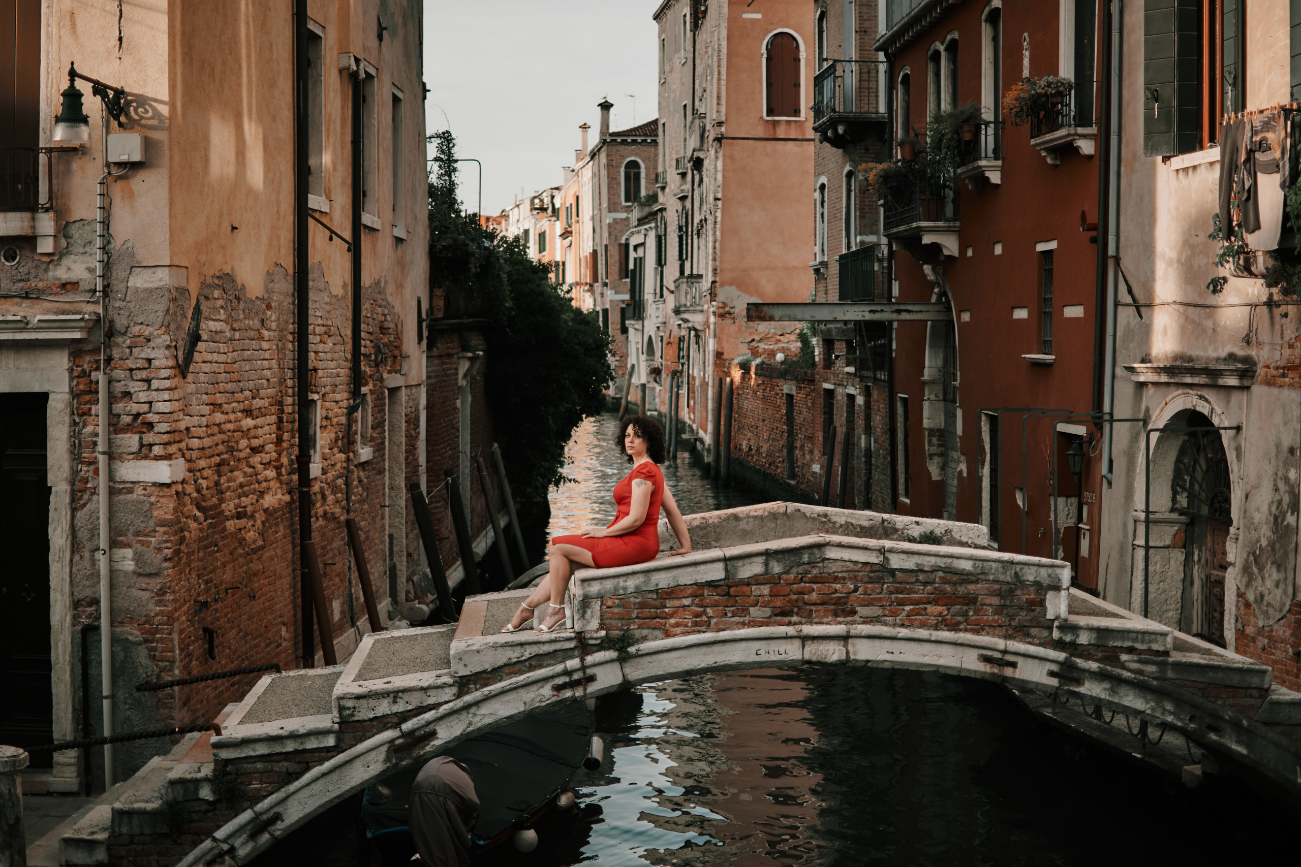 A scenic view of Ponte Chiodo, a historic bridge in Venice, with a woman in a striking red dress standing on it. The bridge is devoid of railings and has a charming rustic appeal. The woman stands gracefully, her flowing red dress contrasting beautifully against the muted tones of the surrounding architecture. The tranquil canal beneath the bridge adds to the serenity of the scene. This image captures the elegance and allure of Venice, with the woman in red adding a touch of glamour and allure to the composition. A captivating moment on Ponte Chiodo that encapsulates the timeless charm of Venice.