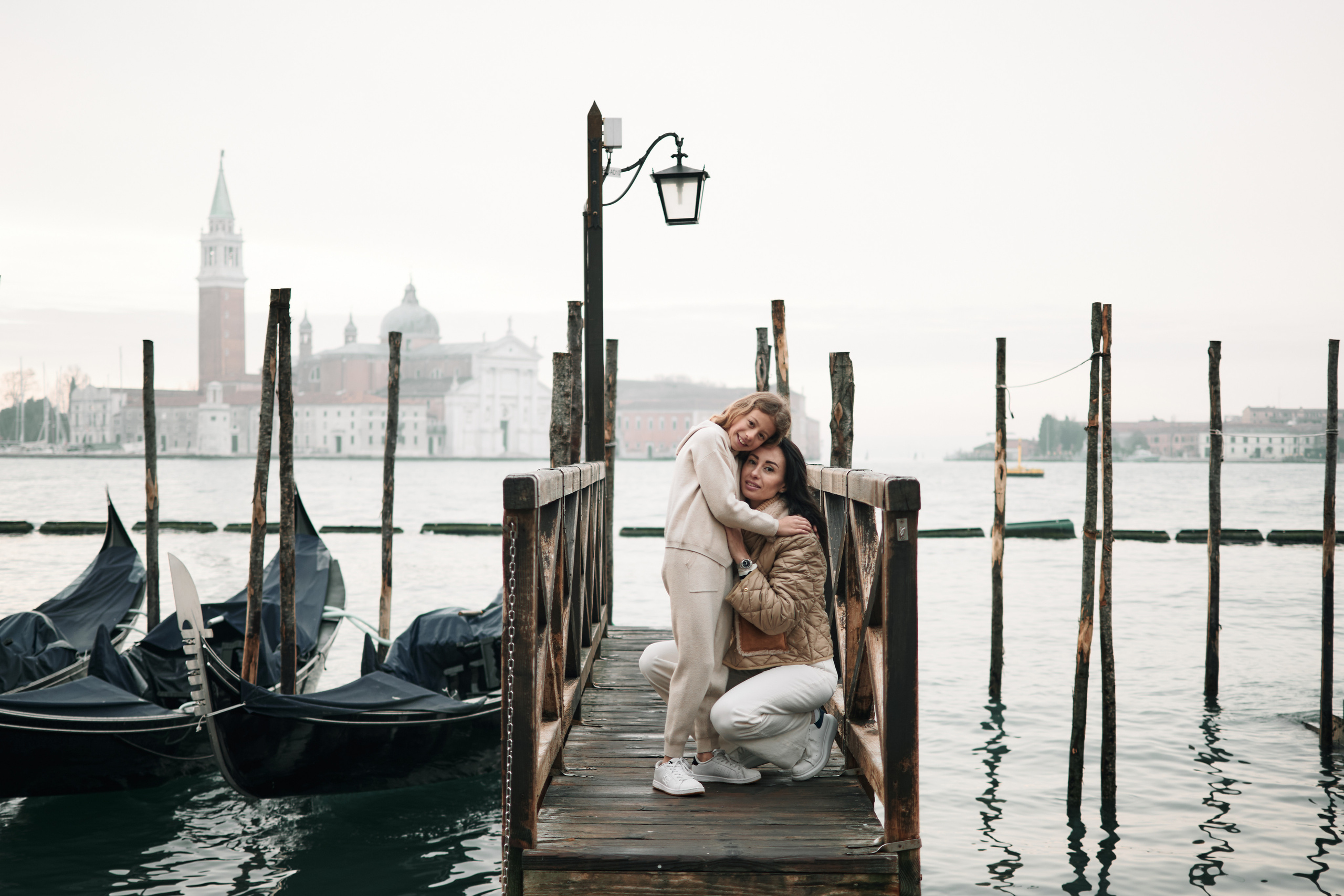 In the heart of Venice, a mother and child share a moment of quiet contemplation on San Marco square. The city's beauty is reflected in their peaceful expressions. A portrait of a mother and daughter on San Marco square in Venice, Italy, taken on a cloudy day. The mother stands behind her daughter, with her arms around her and a warm smile on her face. The daughter looks up at her mother, also smiling, with the grand San Marco square in the background. The cloudy sky adds a moody, atmospheric feel to the photo, contrasting with the bright colors of the buildings and the warm connection between the two subjects