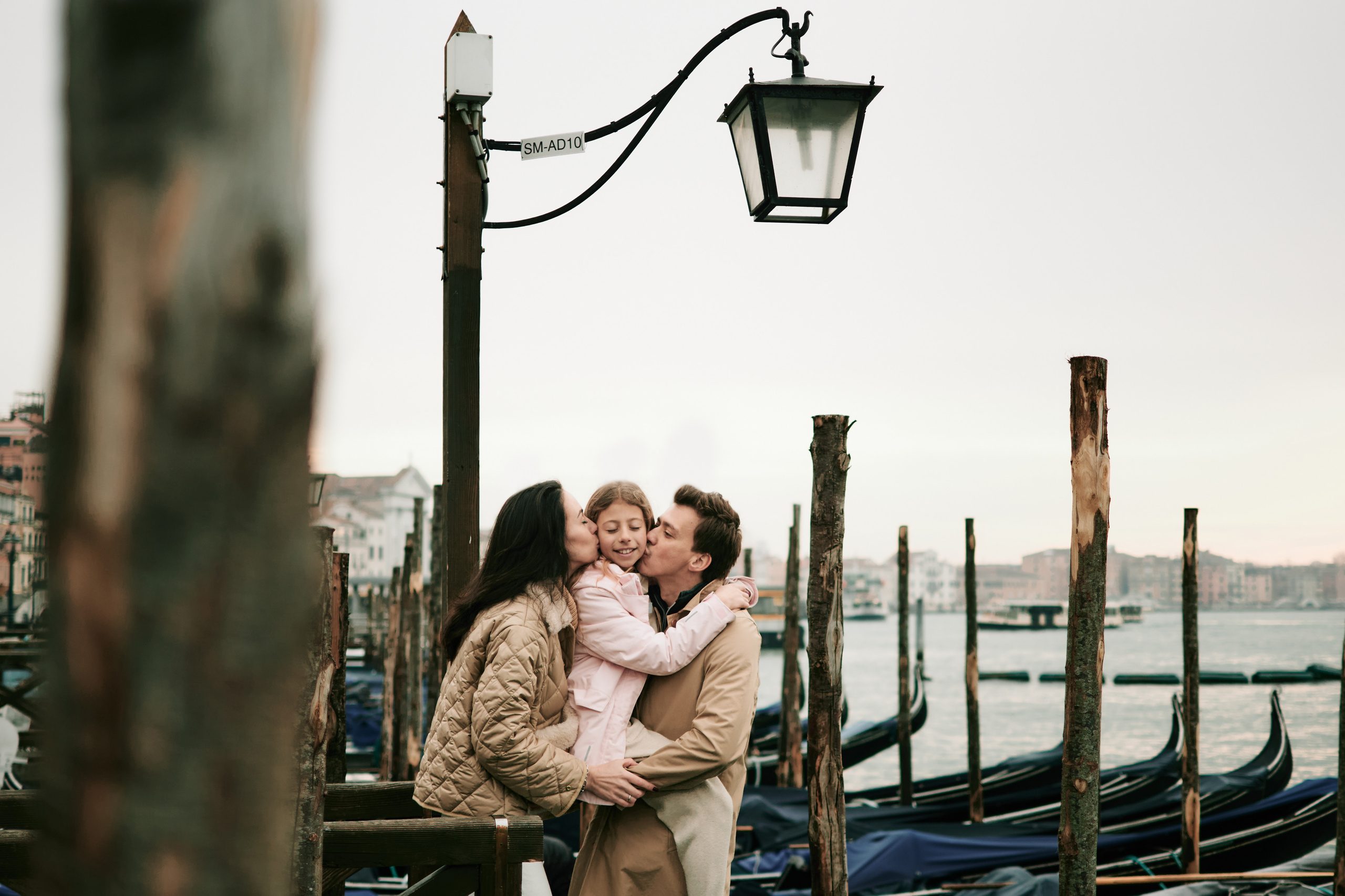 A joyful family stands together on the bustling cobblestone square of San Marco in Venice.  The warm glow of the sun casts a golden light over the scene, highlighting the family's smiling faces and the colorful buildings that line the square.