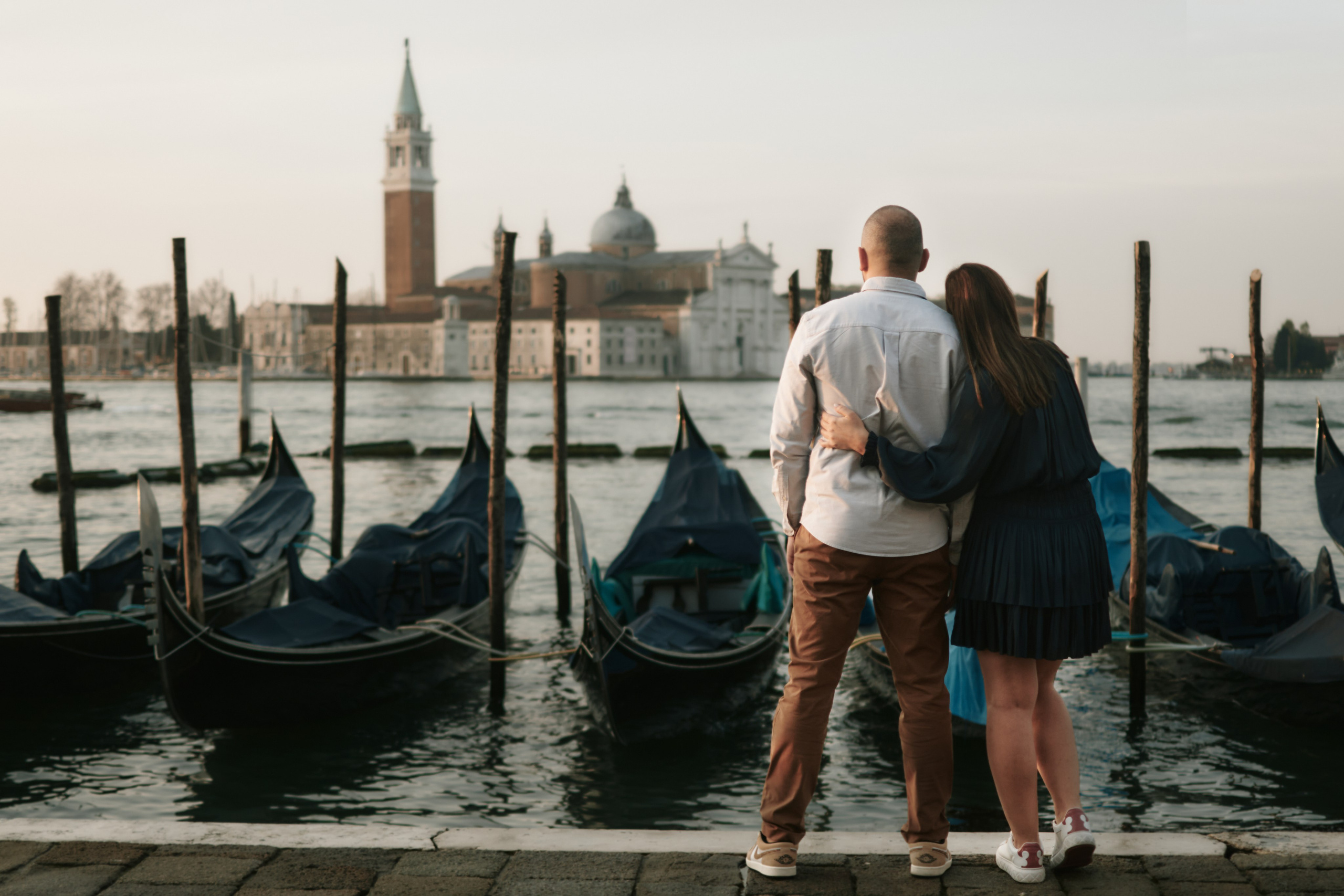 Portrait of a young woman in a black dress standing on San Marco Square, Venice, with a breathtaking panoramic view of the Laguna in the background. The woman has long brown hair styled in loose waves, and she is looking dreamily towards the view. The square is bustling with people, and the stunning architecture of the surrounding buildings adds to the romantic atmosphere of the location
