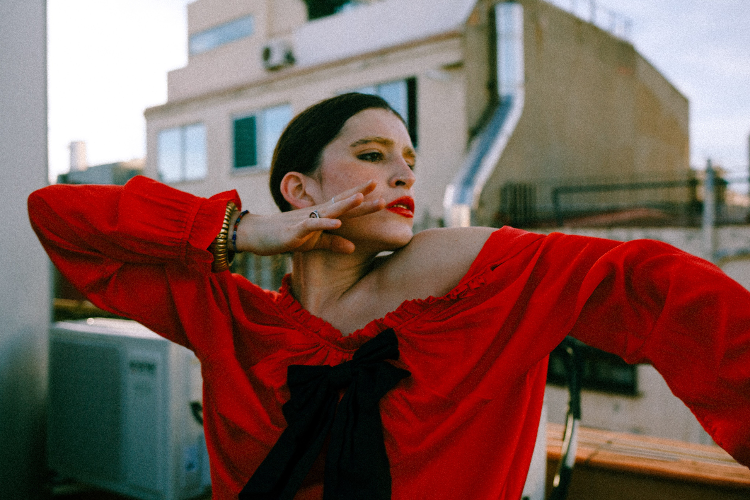 Mujer con blusa roja bailando pensativamente en una azotea con edificios de la ciudad al fondo
