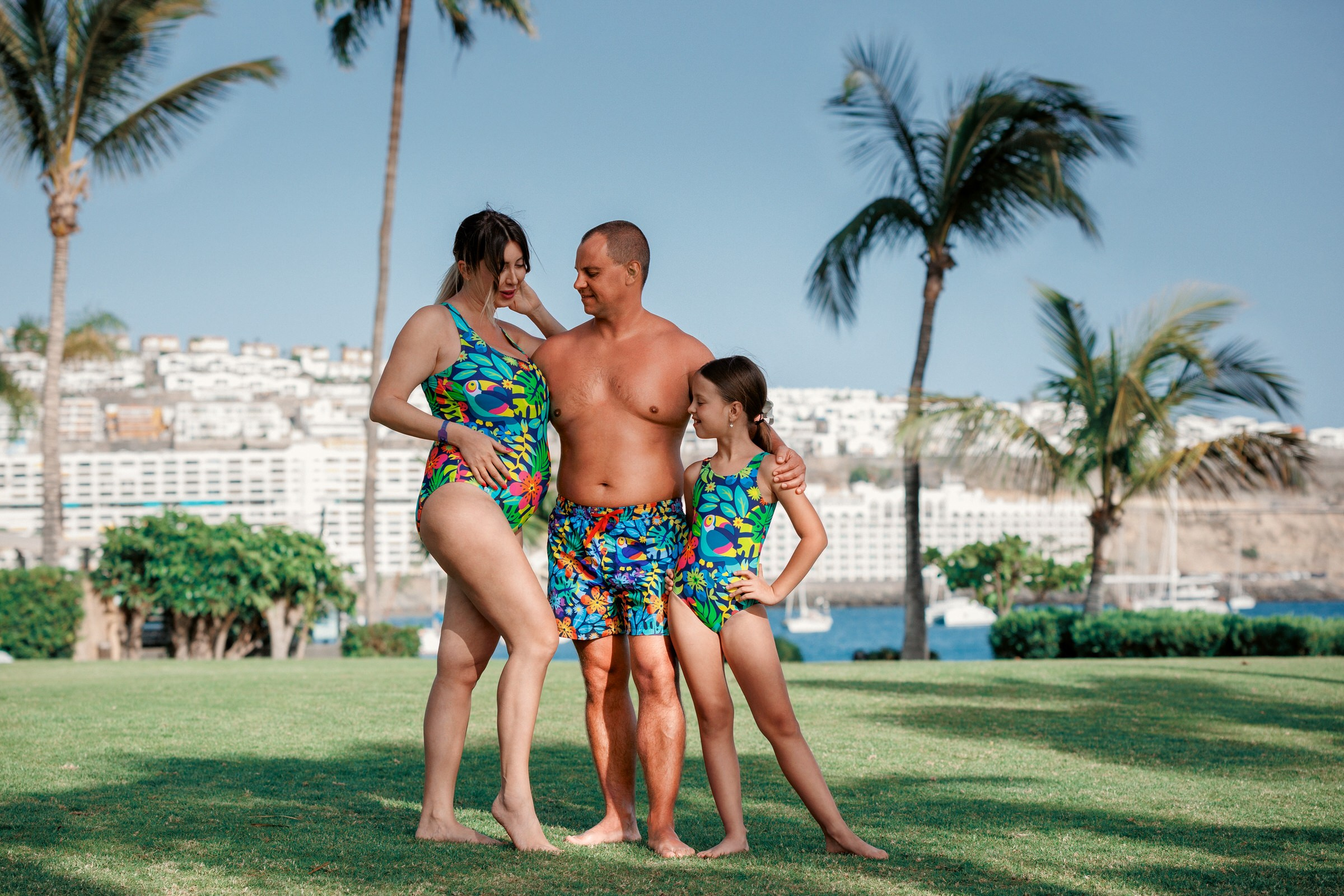 Plage d'Anfi Gran Canaria Une famille de trois personnes portant des maillots de bain tropicaux assortis se tient ensemble sur l'herbe avec des palmiers et des bâtiments en arrière-plan.