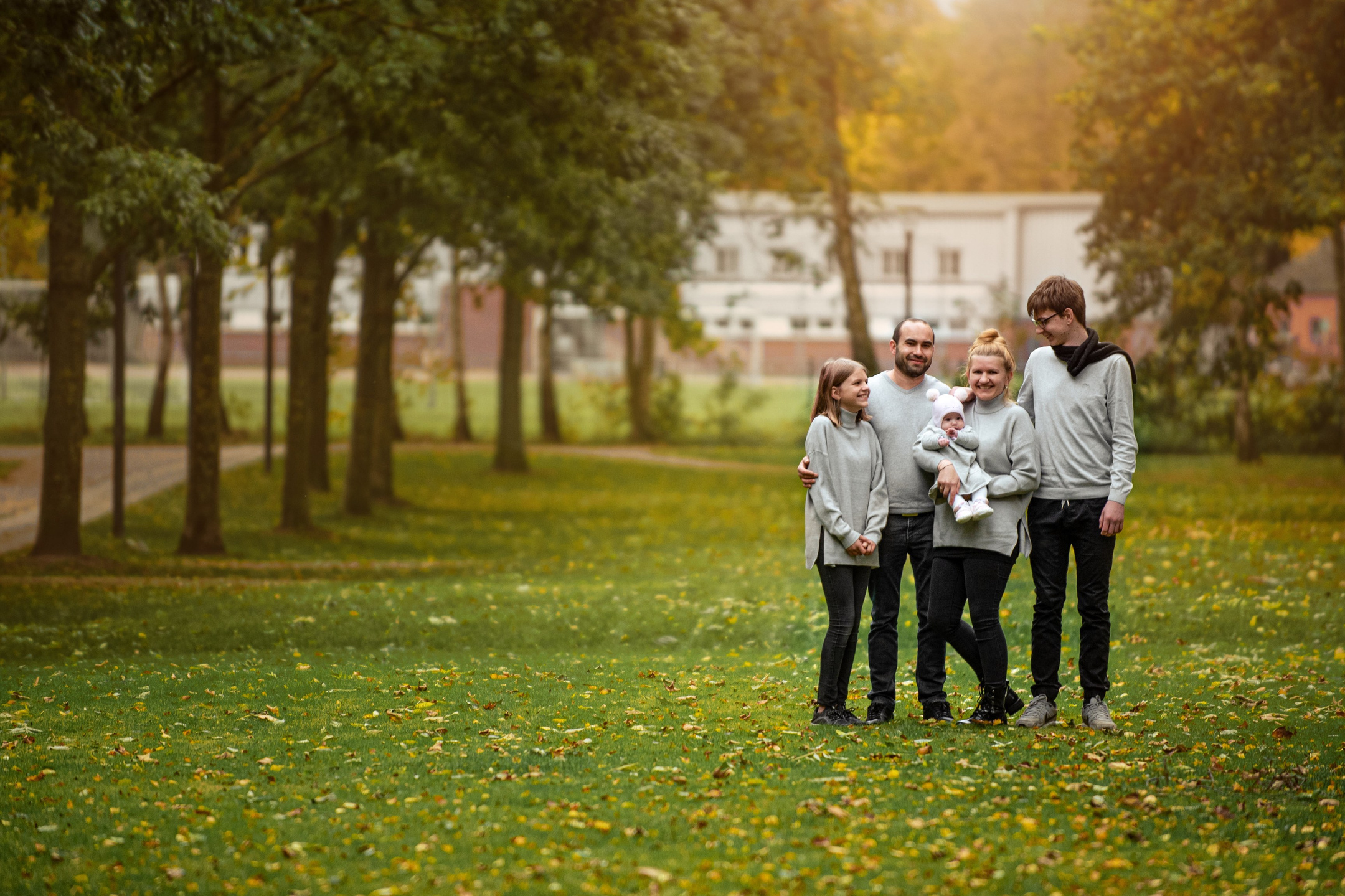 Familie steht zusammen auf einer grünen Wiese in einem Park, umgeben von Bäumen. Alle tragen graue Pullover, und eine Person hält ein Baby im Arm. Die Stimmung ist freundlich und entspannt, mit einem herbstlichen Hintergrund aus buntem Laub und Gebäuden in der Ferne
