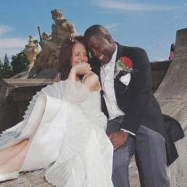Red-haired bride holding her flowing veil aloft smiles seductively to her bouquet holding groom as he leans back against a railing in appreciation at the Vrtba Garden in Prague. 