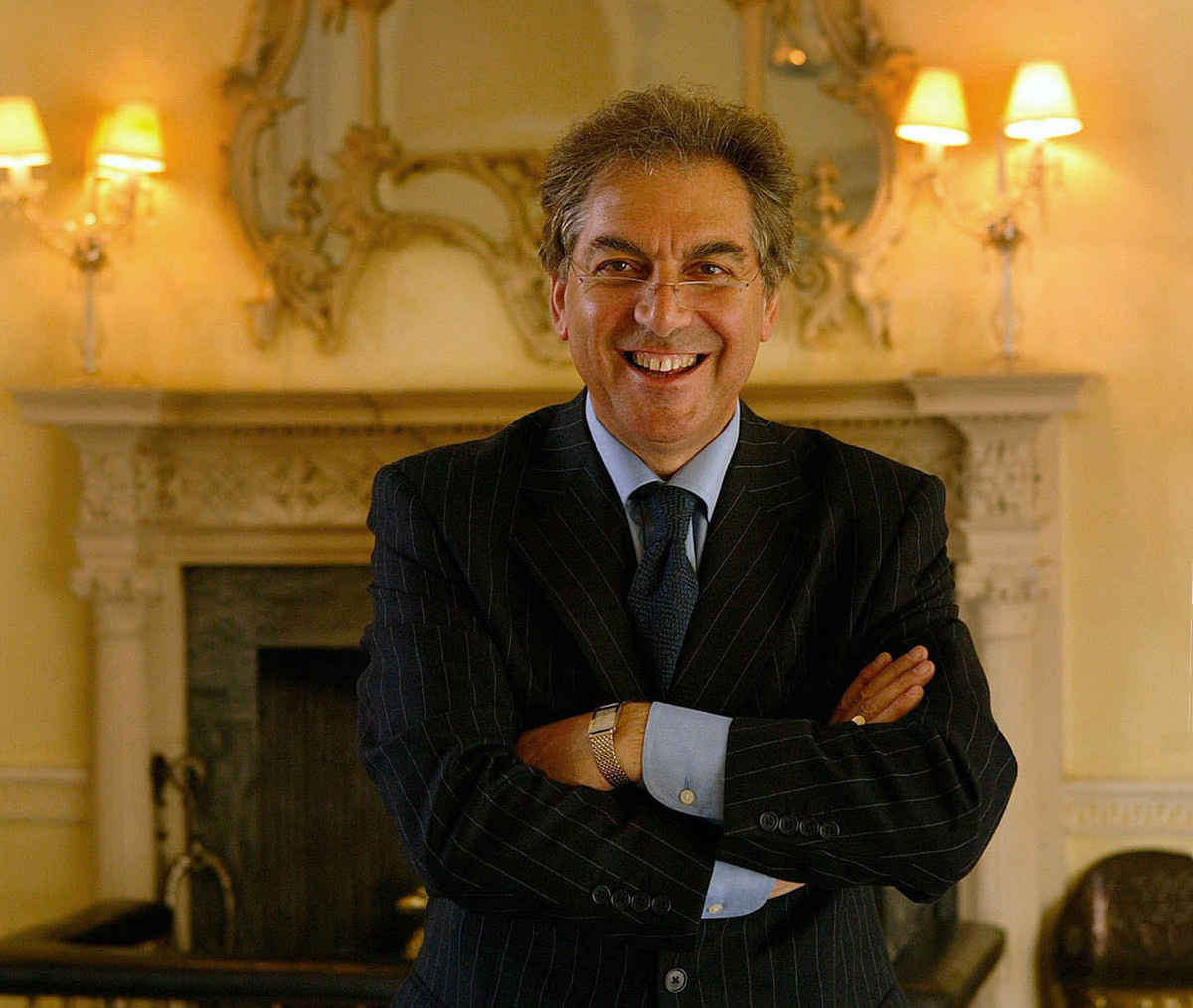 An elegantly dressed older man poses for a executive portrait near a fireplace in a 17th-century lodge.