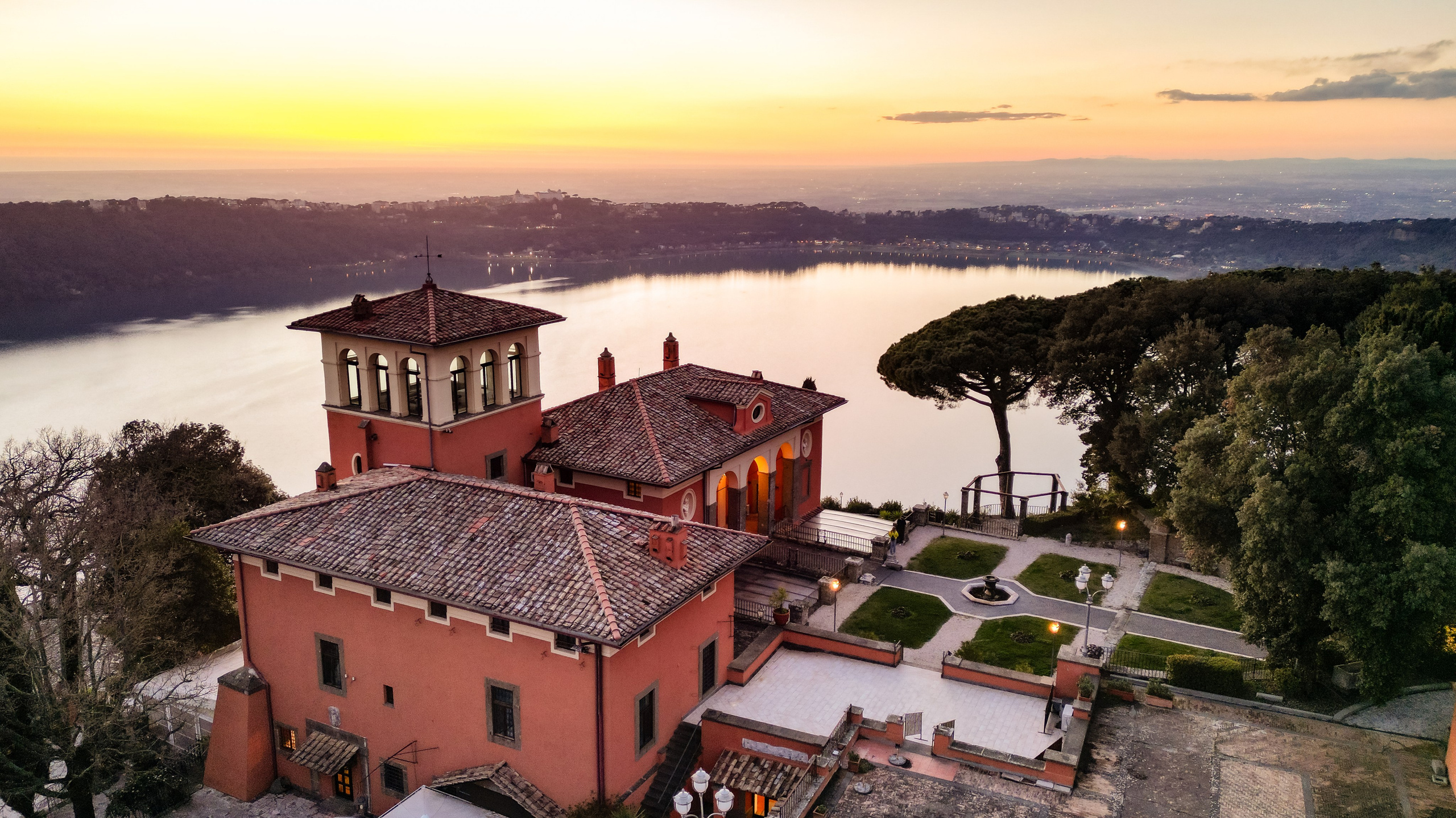 Vista panoramica al tramonto della maestosa Villa del Cardinale che si affaccia sul Lago Albano, con il suo belvedere e i caratteristici tetti rossi che si stagliano contro il cielo infuocato.