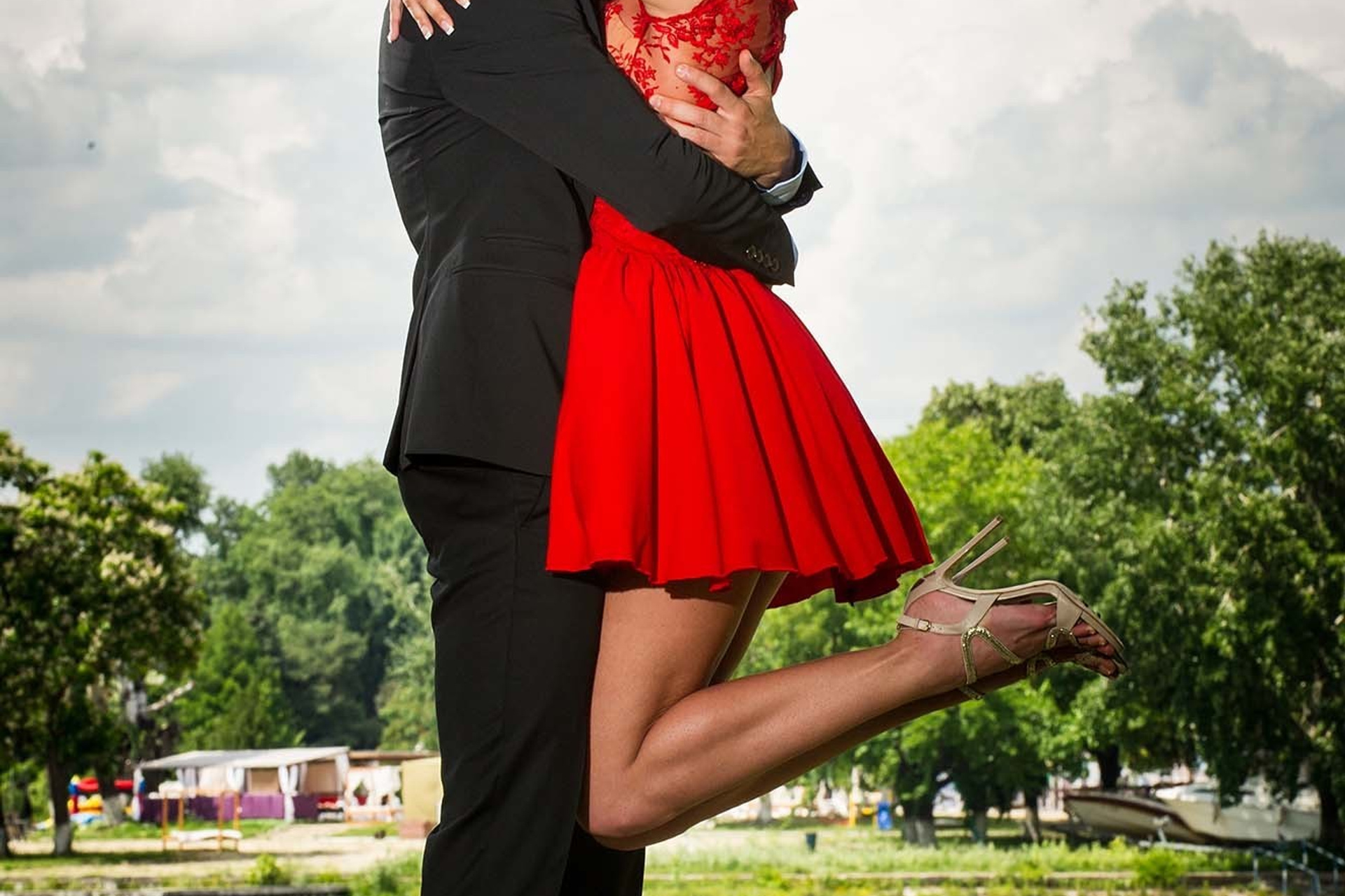 Man lifting woman in red dress while kissing near riverside under cloudy sky.
