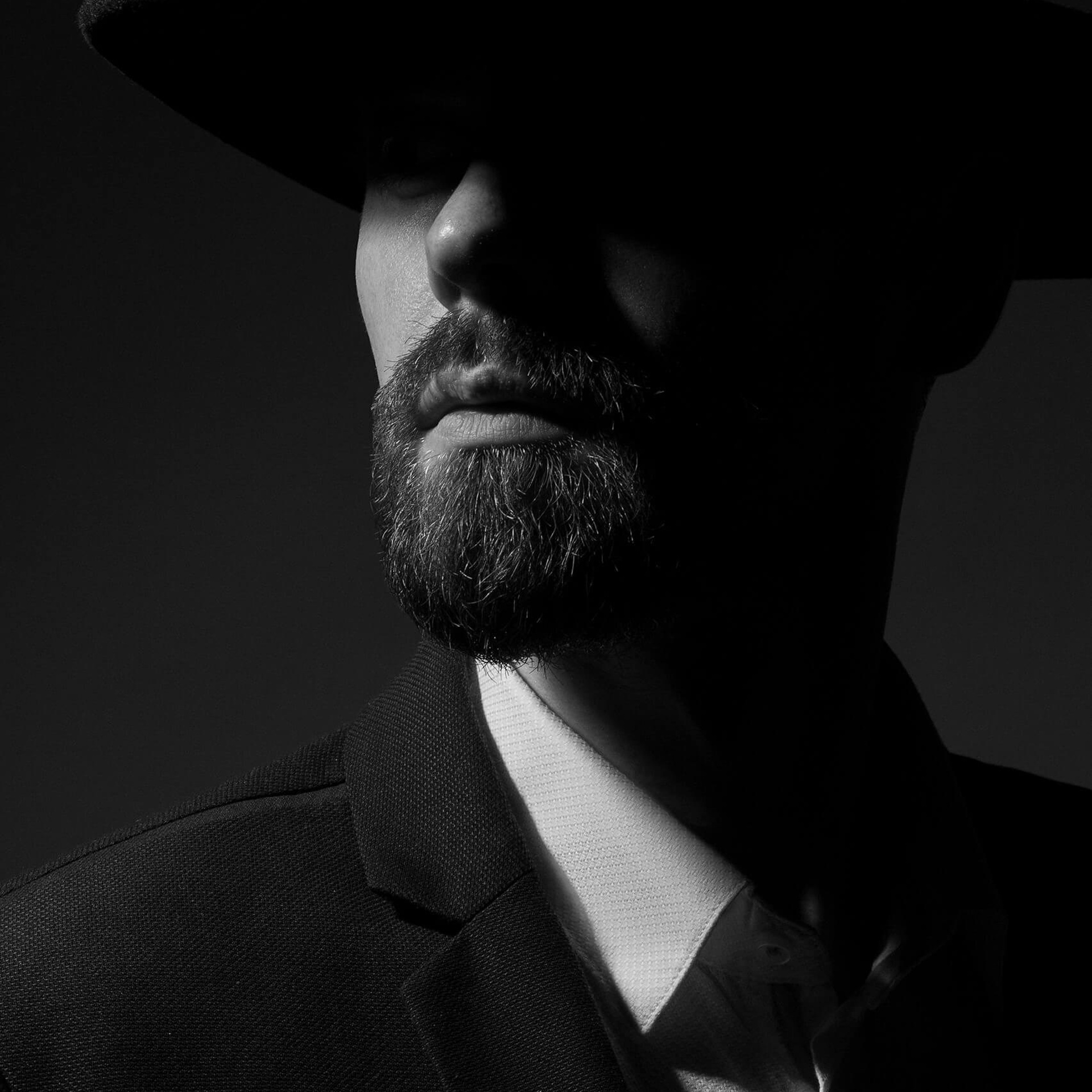 A mysterious and dramatic low-key black and white portrait, featuring a man in a hat with his face artfully shadowed, emphasizing the texture of his beard and the intensity of his gaze