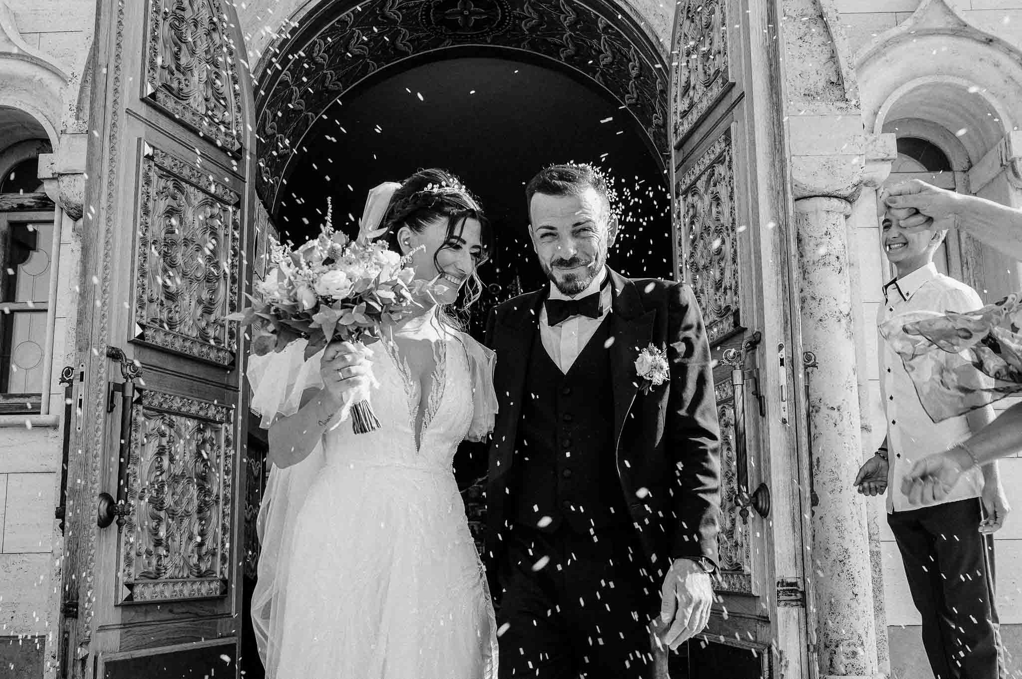Sposi escono sorridenti dalla chiesa sotto una pioggia di riso, lui in un elegante smoking e lei in un abito da sposa con bouquet, con un'antica porta decorata che fa da sfondo a questo momento classico di festa nuziale.